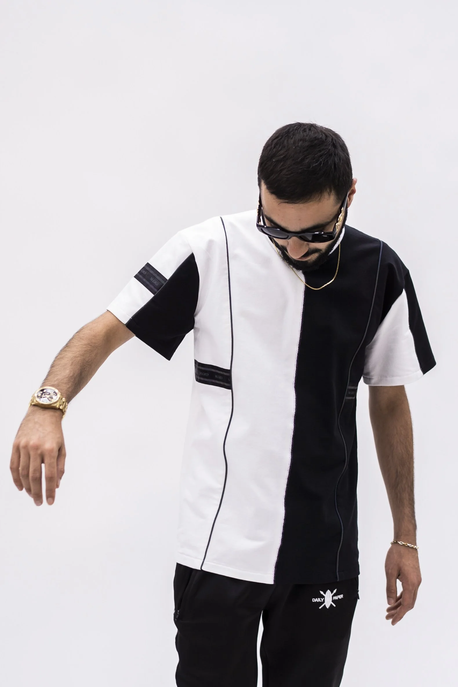 Homme portant une chemise noire et blanche, lunettes de soleil, et bracelets, posé contre un fond blanc.