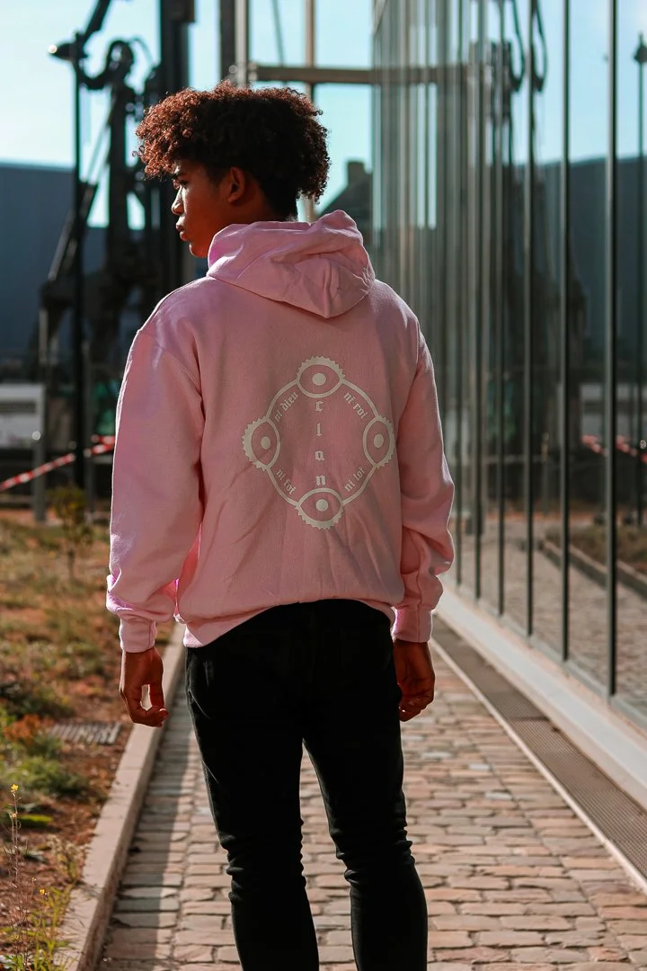 Une jeune femme à cheveux bouclés portant un sweat rose, marchant sur un chemin pavé à côté d'une cloison vitrée, en plein air avec un ciel ensoleillé.