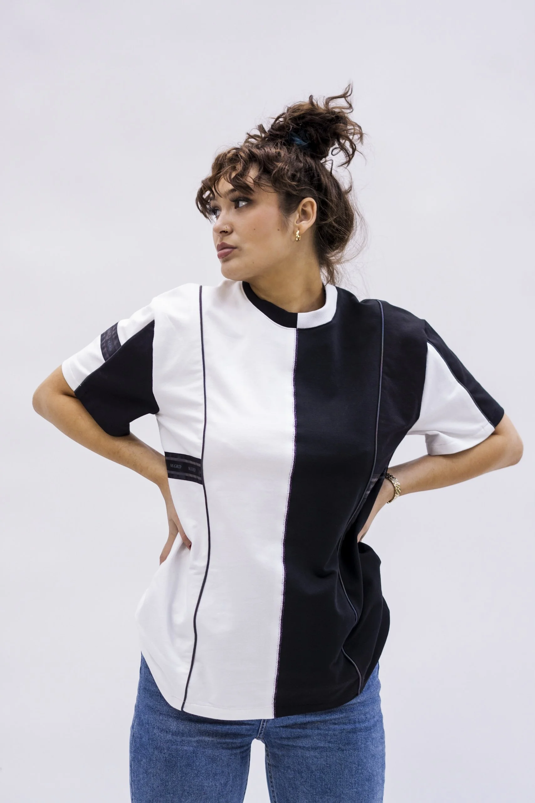 Une femme avec des cheveux bouclés en haut, portant un t-shirt à moitié blanc et noir, avec des mains sur les hanches, devant un mur blanc.