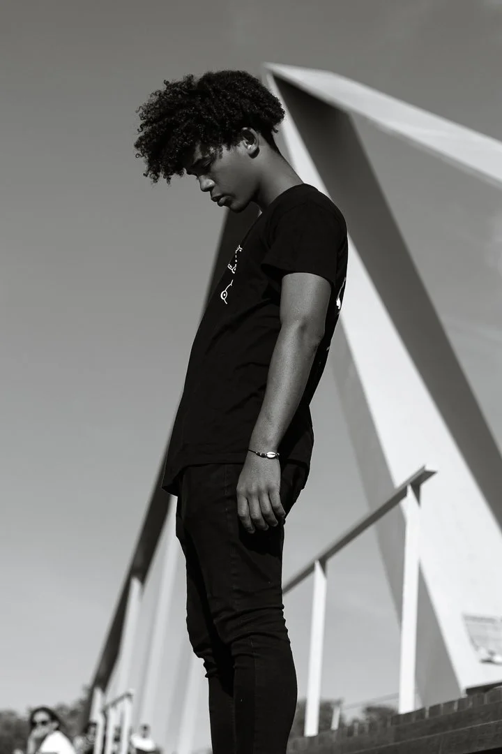 Un jeune homme noir avec des cheveux bouclés portant un t-shirt noir, regardant vers le bas, en extérieur avec une architecture moderne en arrière-plan.