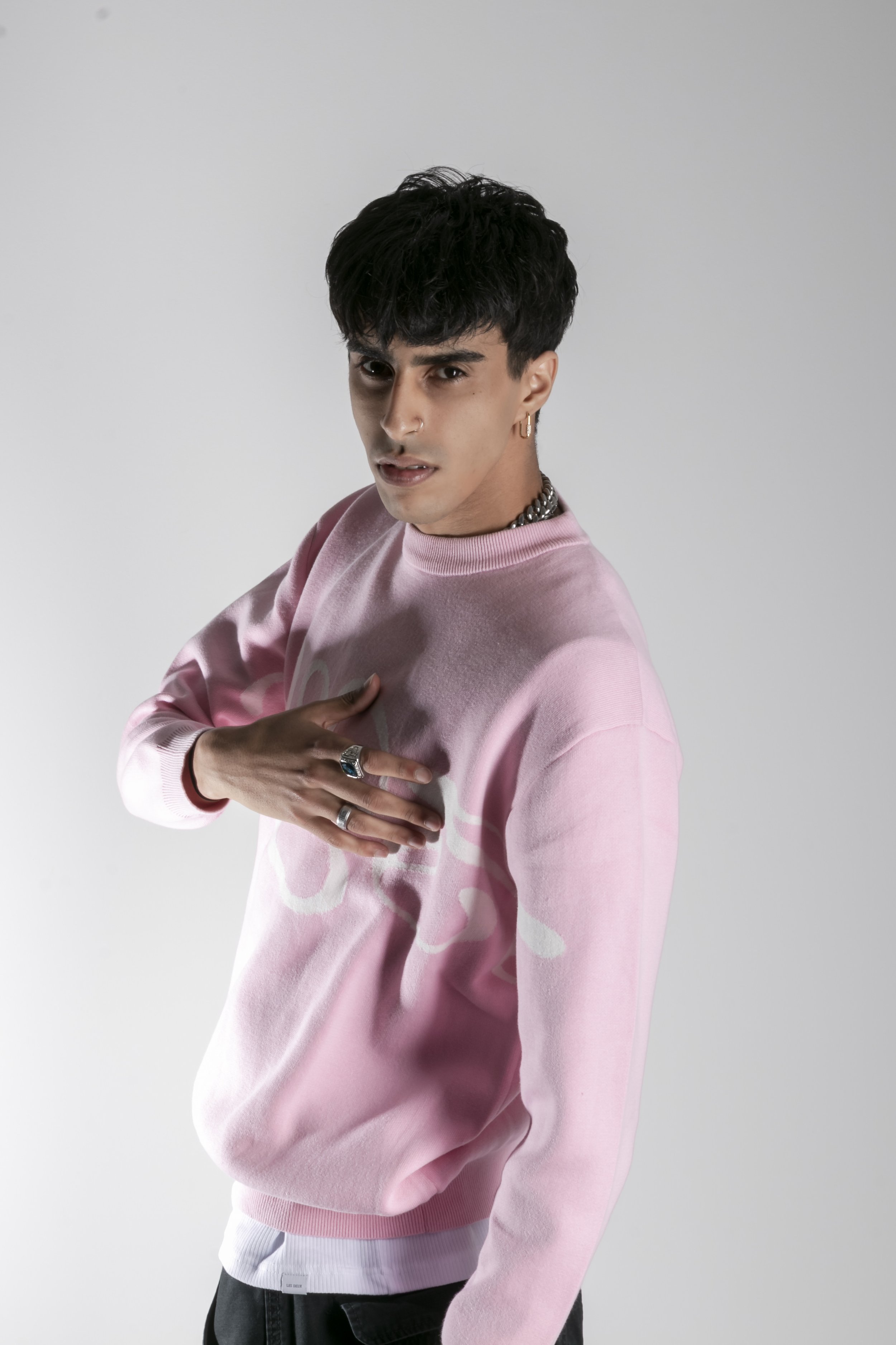 Jeune homme portant un sweat rose et des bijoux, posant avec une main sur la poitrine, regardant l'objectif de manière expressive, contre un fond blanc.