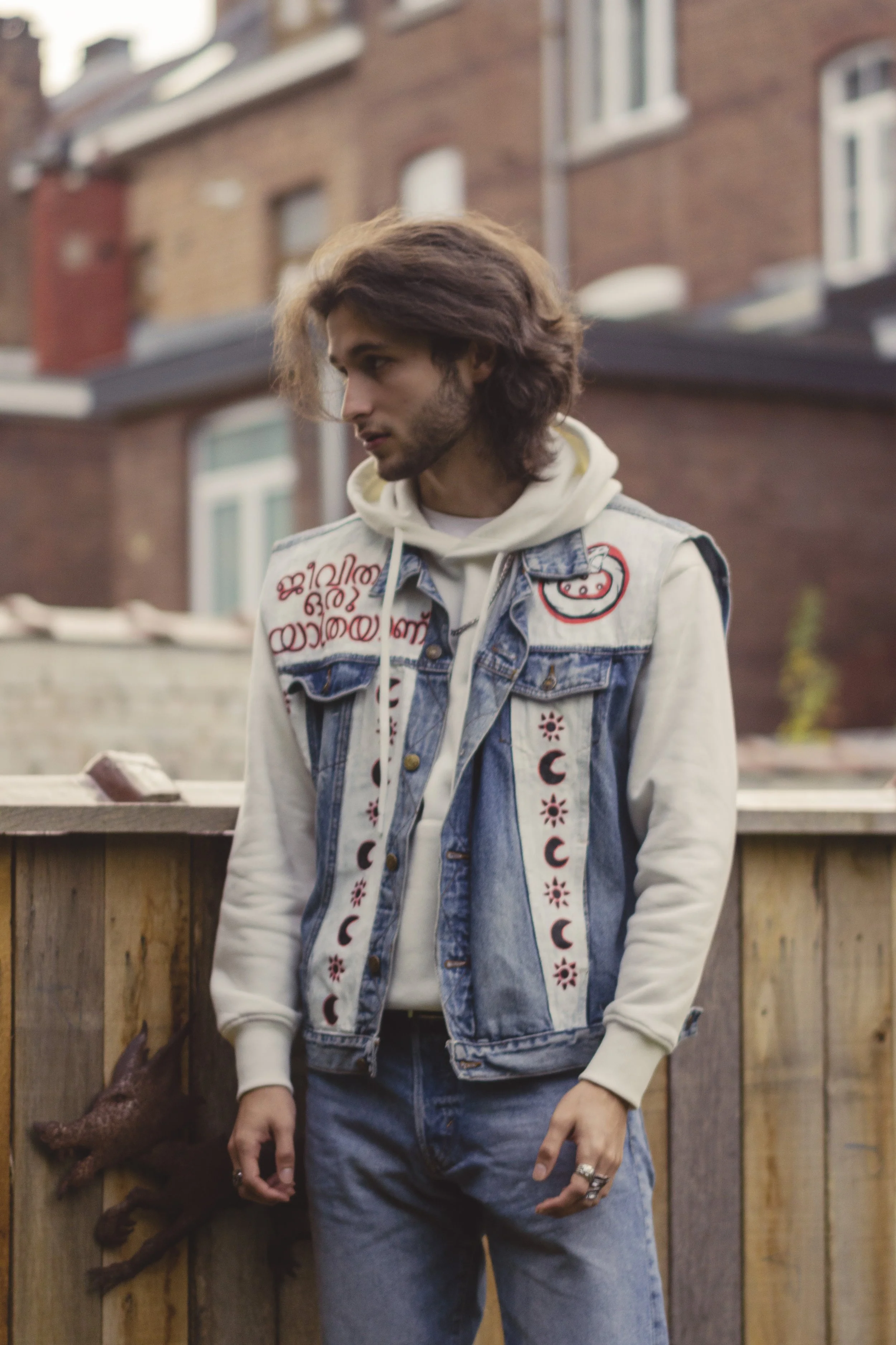 Jeune homme avec des cheveux longs portant une veste en denim décorée de motifs et de texte, avec un sweat à capuche beige, debout à côté d'une clôture en bois dans un quartier résidentiel.