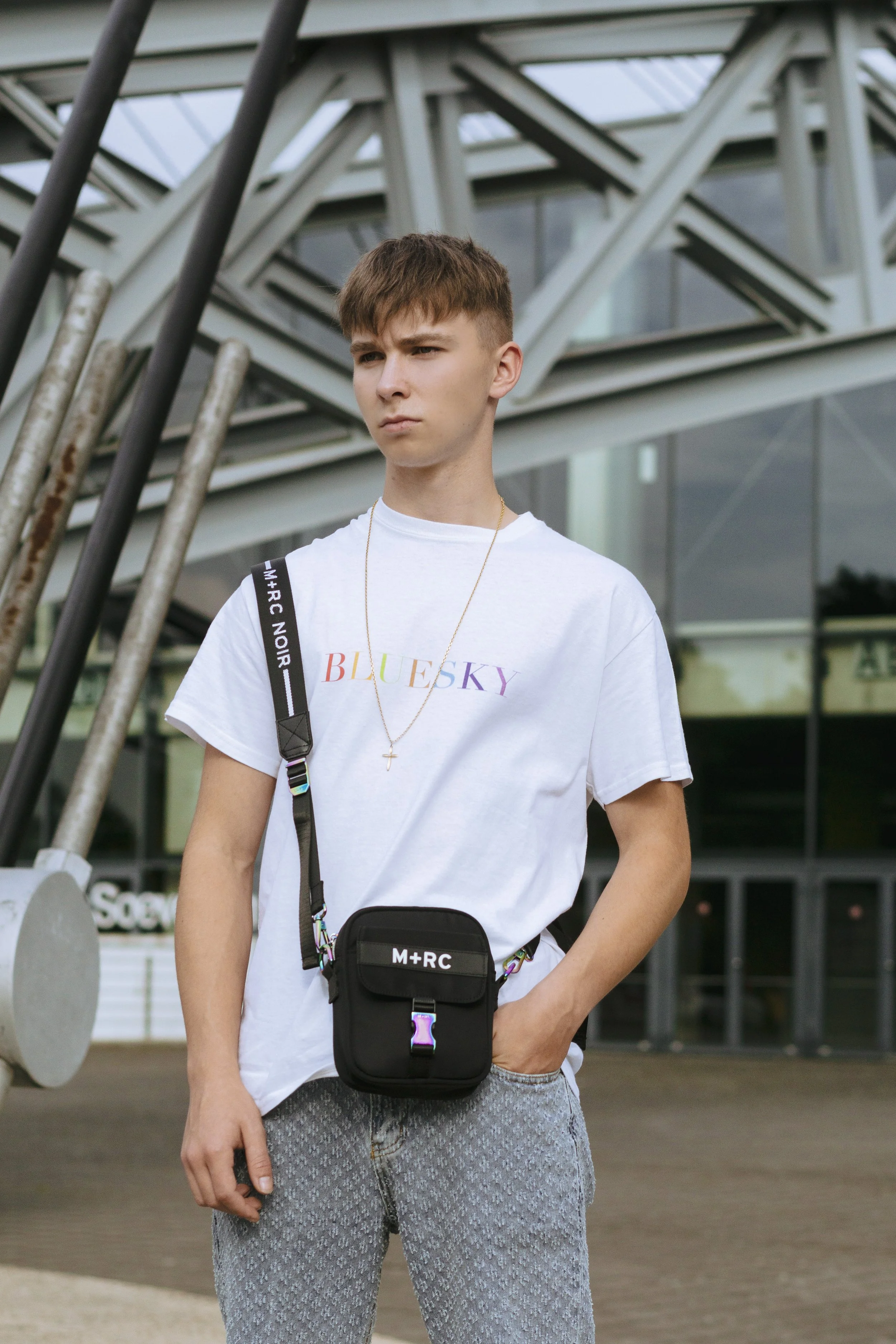 Un jeune homme portant un t-shirt blanc avec l'inscription "BLUESKY" en couleur, un sac à bandoulière noir avec la marque "M+RC", une chaîne avec une croix, et un jean gris, se tient devant une structure métallique moderne.