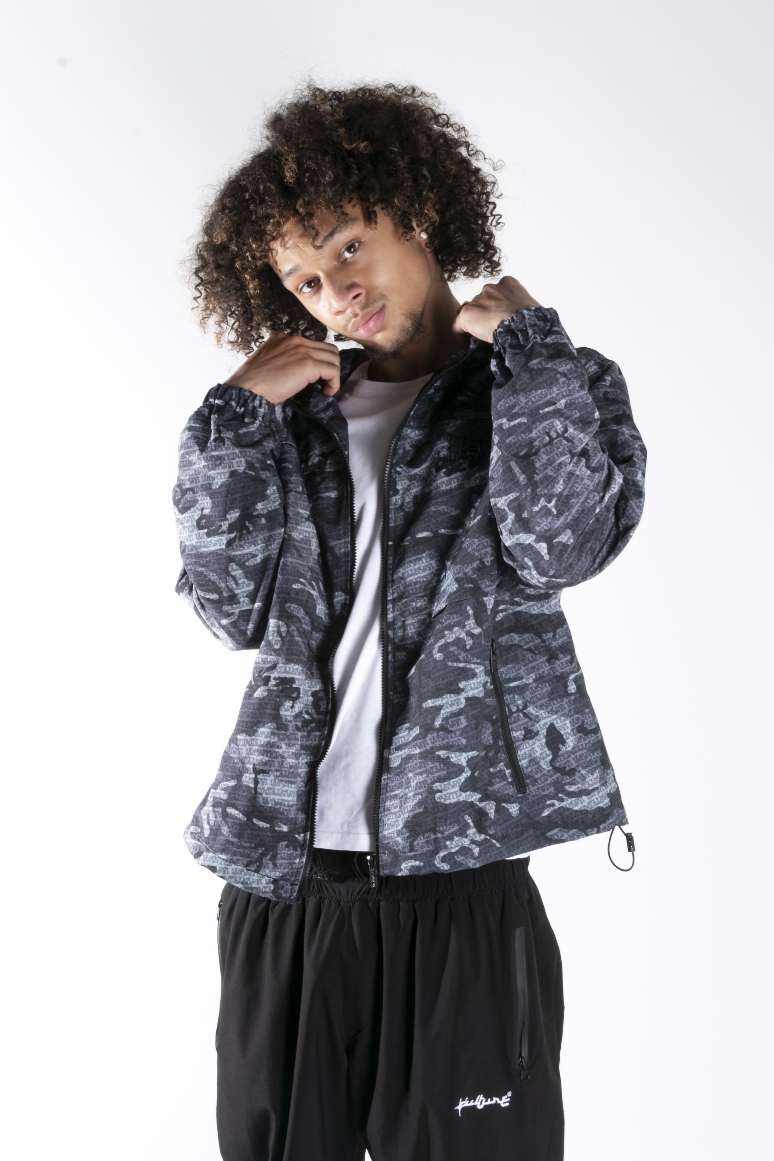Jeune homme avec des cheveux bouclés portant une veste noircie à motif camouflage gris, une chemise blanche et un pantalon de survêtement noir, posé contre un fond blanc.