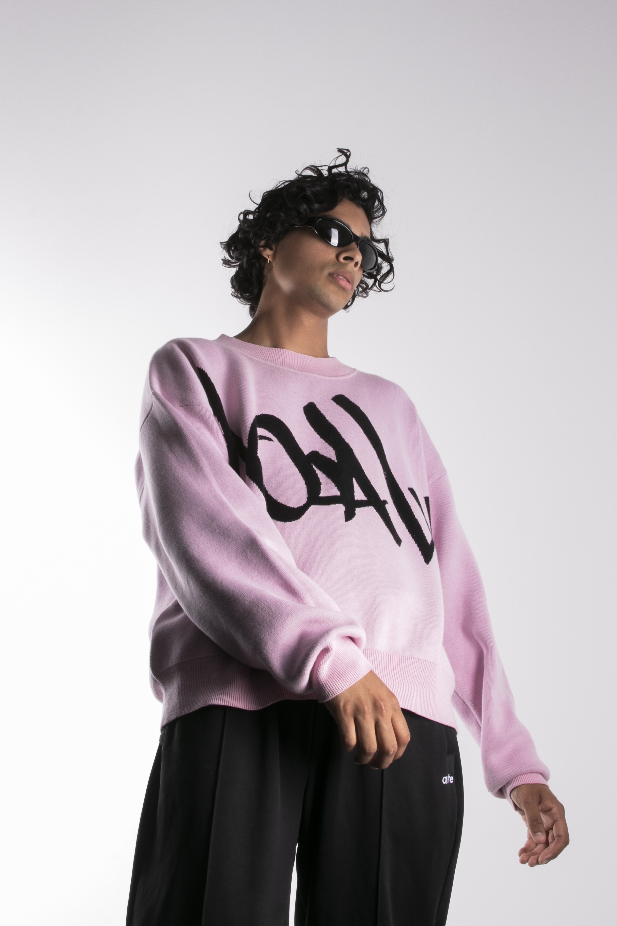 Une femme portant un sweat-shirt rose avec une inscription graphique noire, des lunettes de soleil noires et un pantalon noir, debout contre un fond blanc