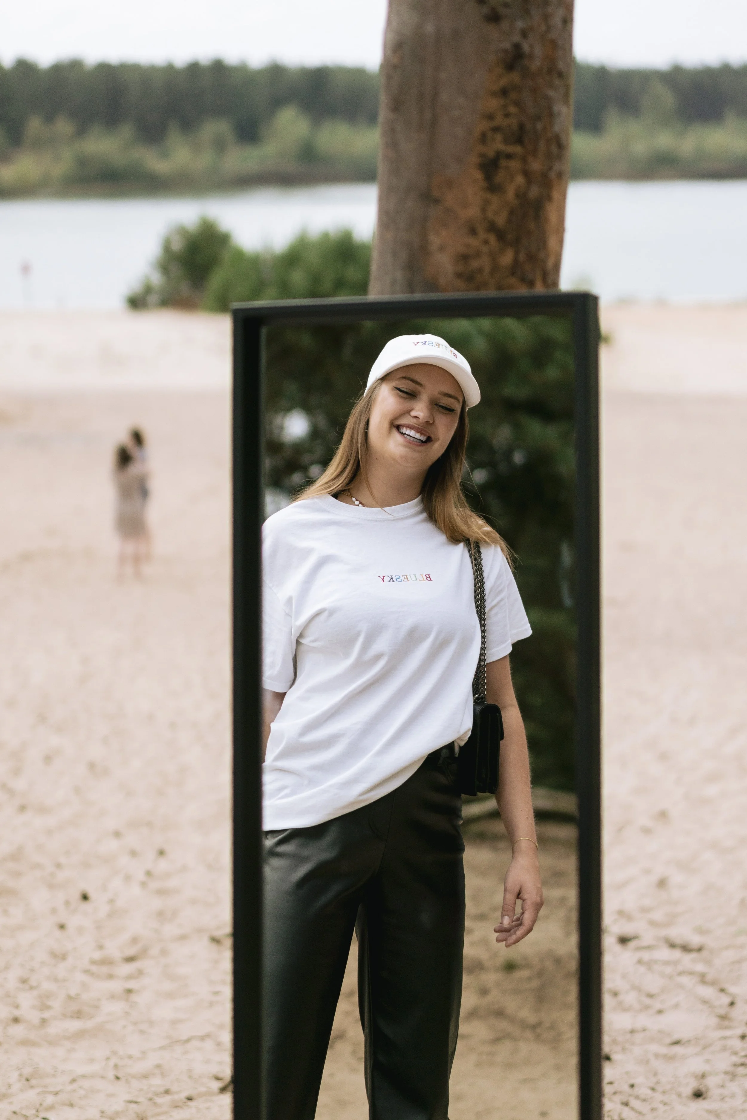 Une jeune femme souriante se regarde dans un miroir placé dans la nature, portant un t-shirt blanc et une casquette blanche.