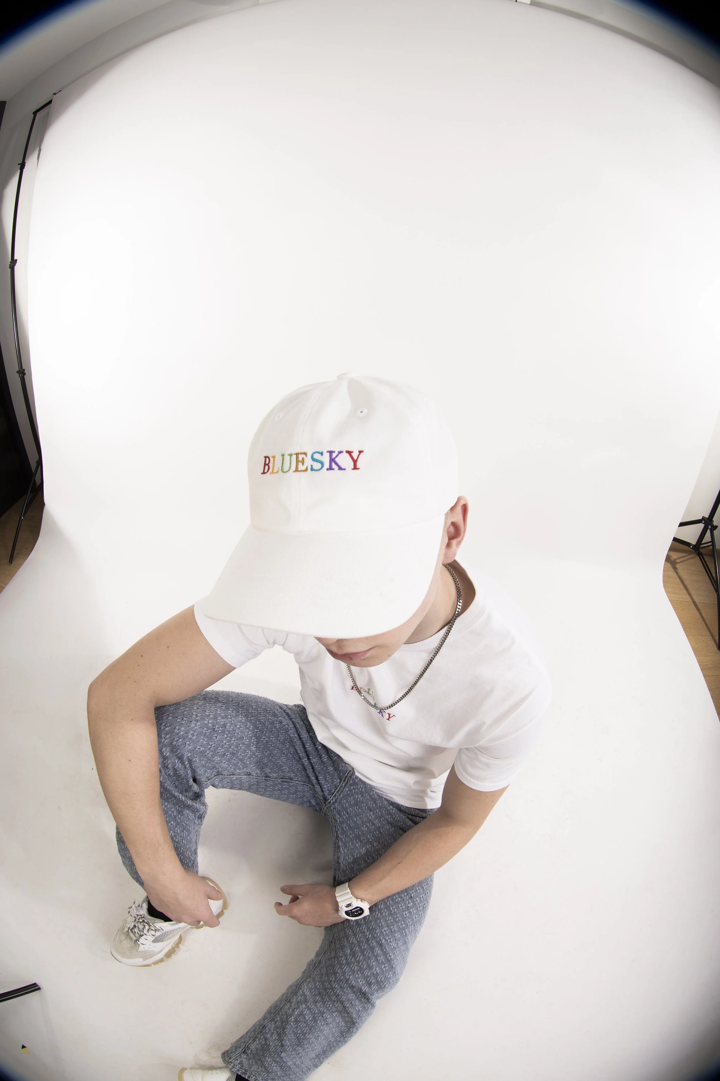 Jeune homme portant une casquette blanche avec l'inscription "BLUESKY" multicolore, une chaîne en or, un bracelet blanc, une t-shirt blanc et un pantalon gris, assis sur un fond blanc en studio. La photo est prise en mode fisheye.