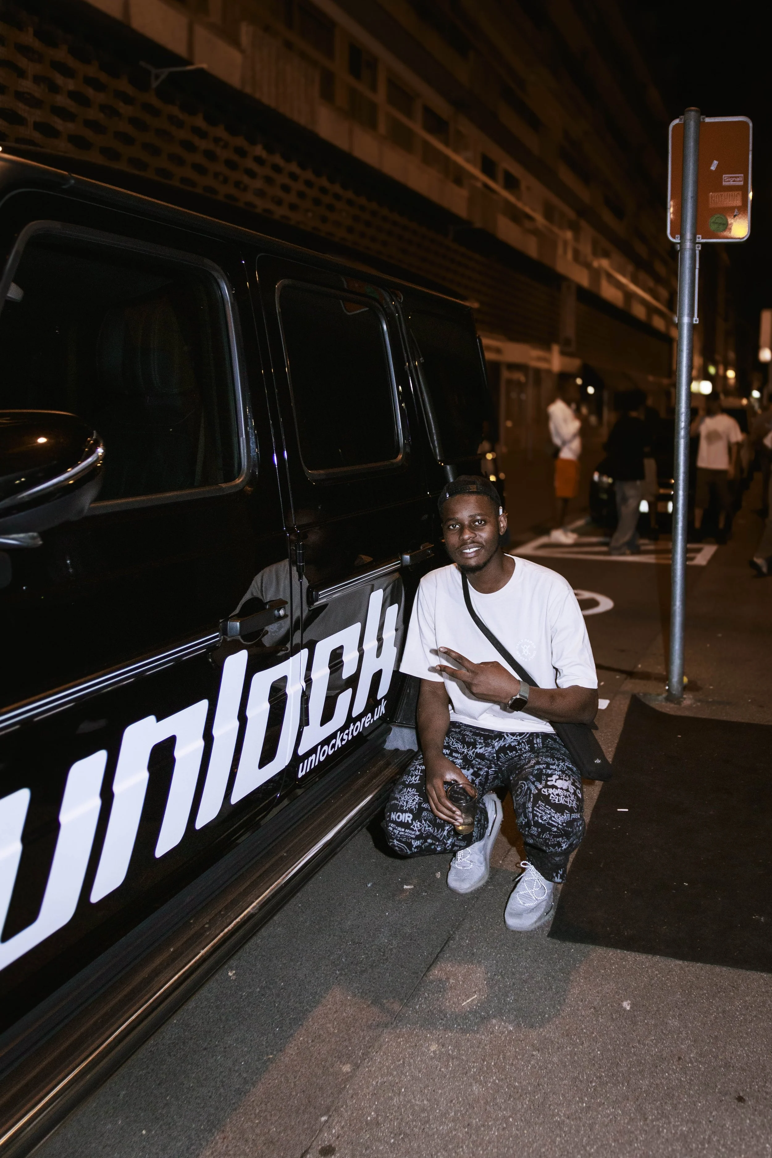 Prodige Mbonzo posé à côté d'un véhicule noir avec un logo 'unlock' visible sur la porte, en pleine rue la nuit.