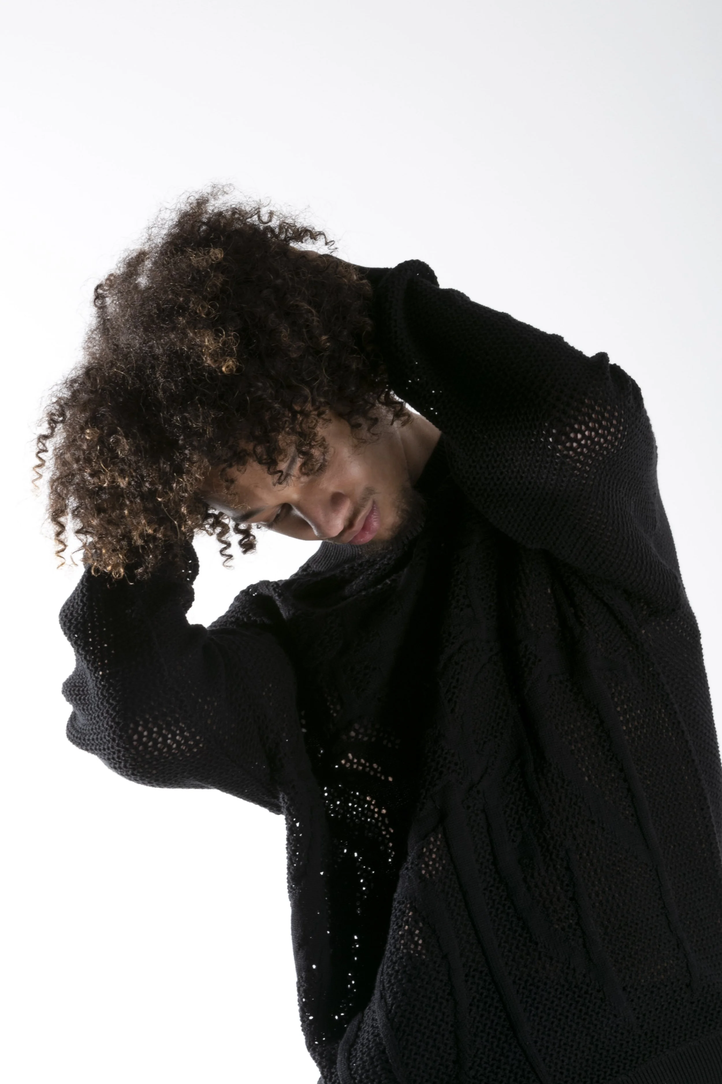 Jeune homme aux cheveux bouclés avec une main derrière la tête, portant un pull noir en tricot avec des trous, en position inclinée.