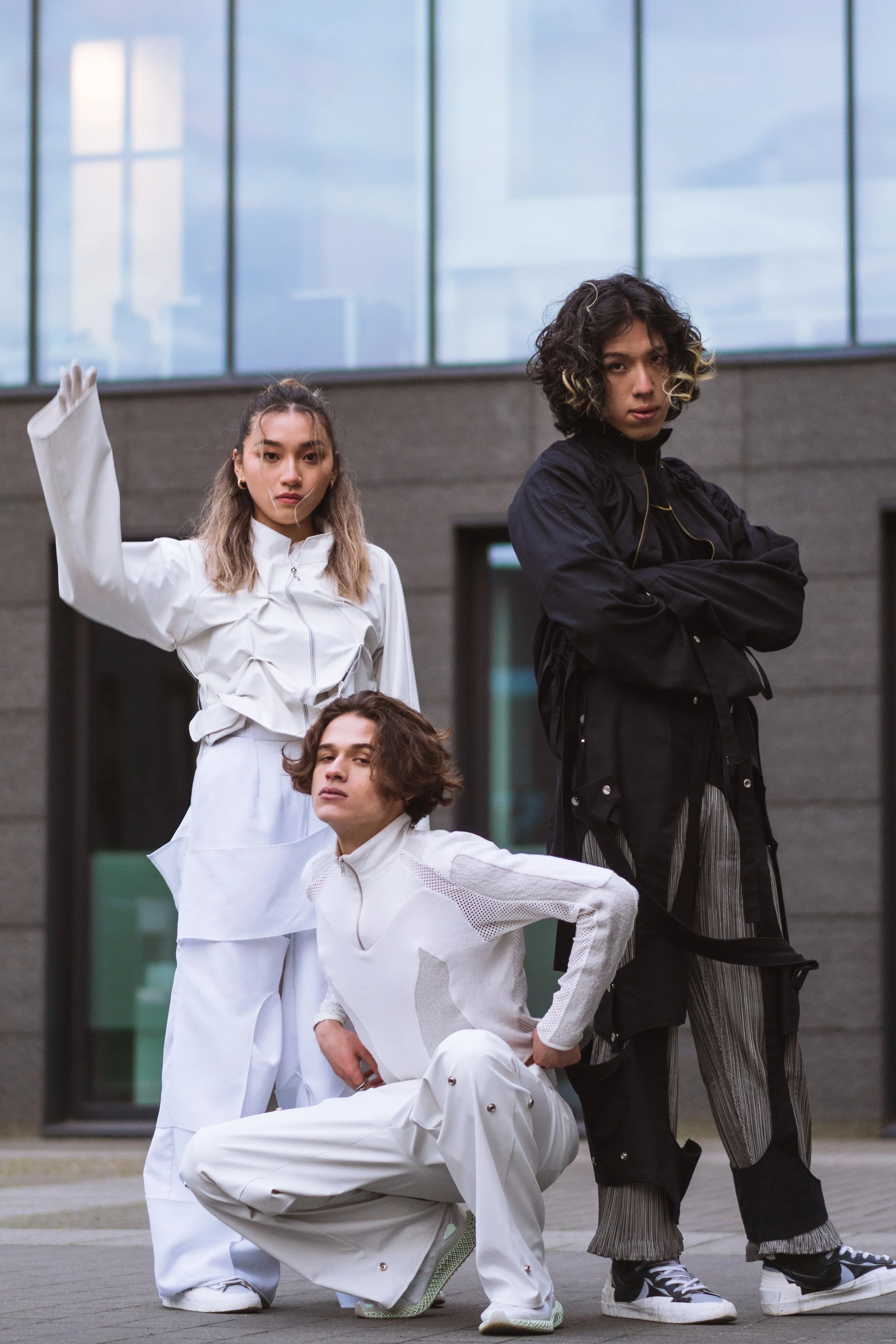 Trois jeunes adultes posant devant un bâtiment moderne avec de grandes fenêtres. La personne à gauche porte un vêtement blanc, celle au centre porte également du blanc, et celle à droite porte un vêtement noir.