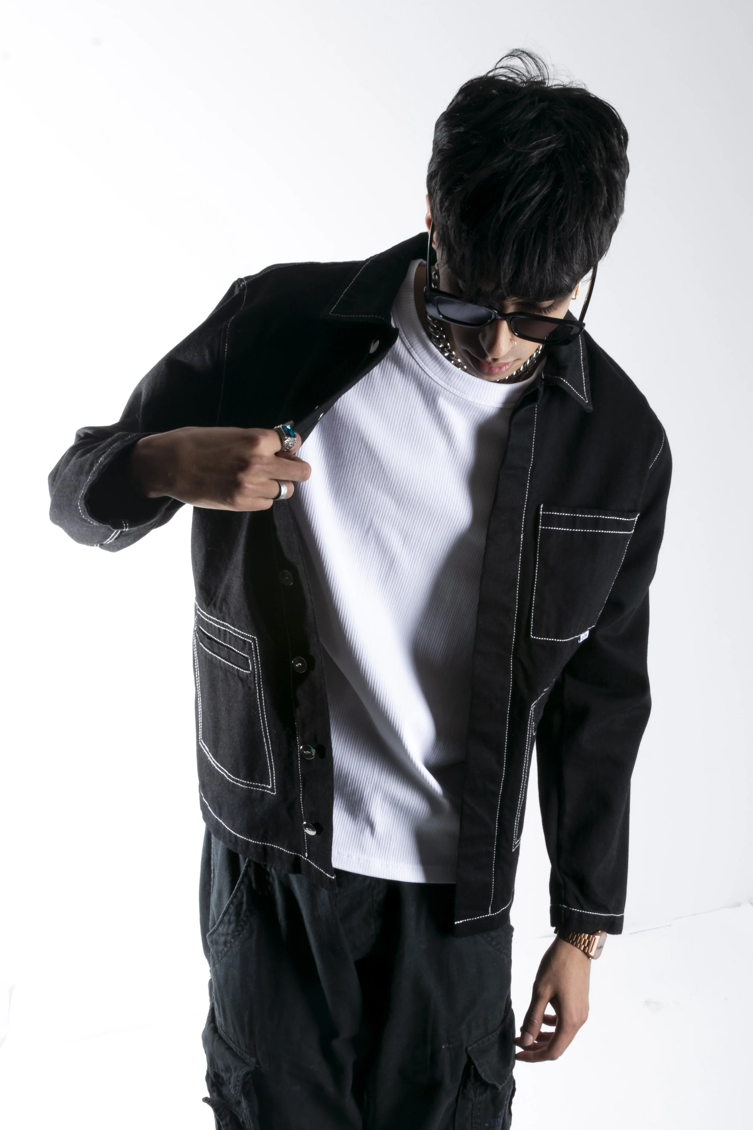 Un homme portant des lunettes de soleil noires, un collier en chaîne, une veste noire à surpiqûres blanches, un t-shirt blanc et un pantalon noir, pose contre un fond blanc.