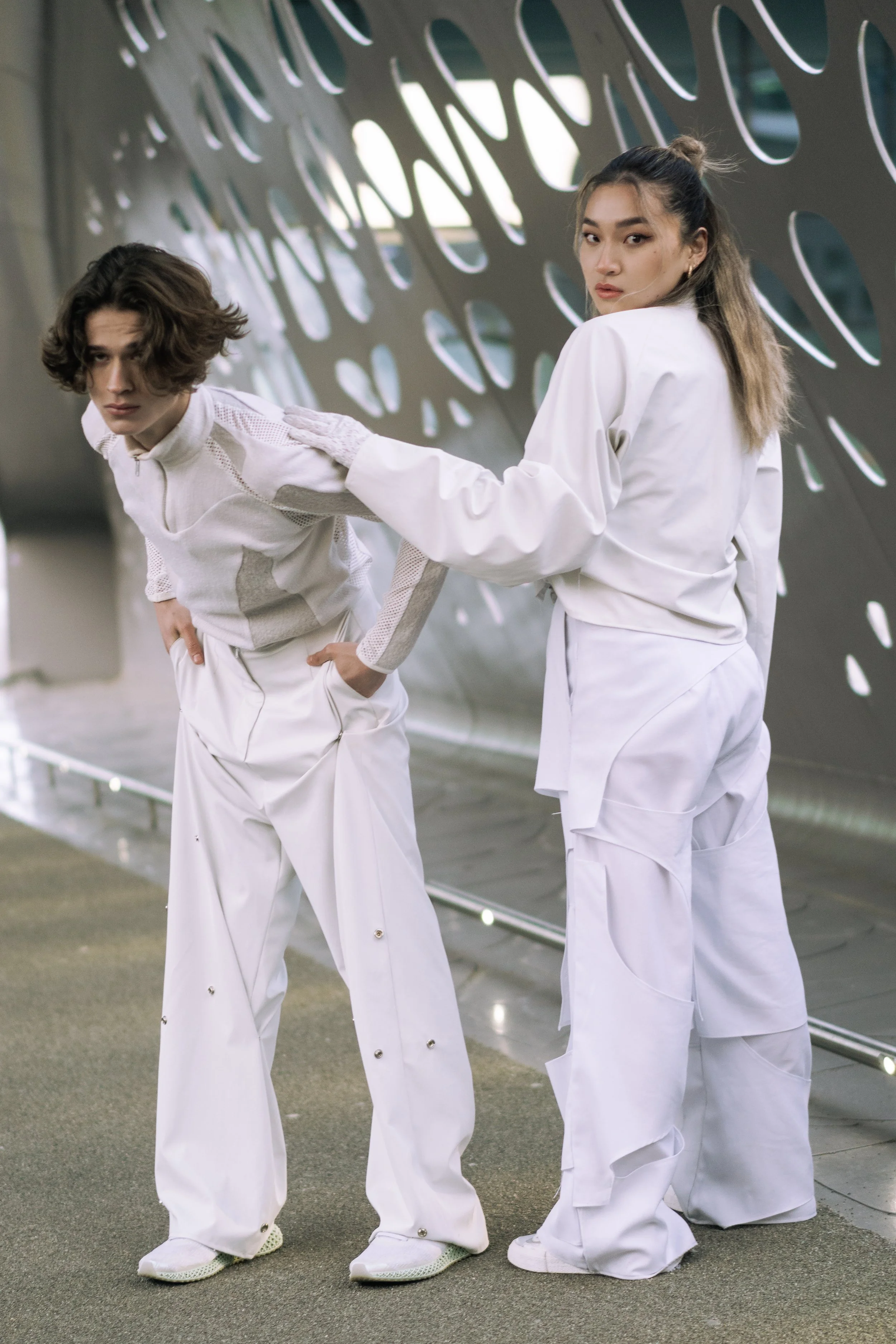 Deux jeunes personnes, un homme et une femme, habillés en vêtements blancs futuristes, se tiennent dans un environnement moderne avec des motifs circulaires en arrière-plan. La femme touche l'épaule de l'homme.