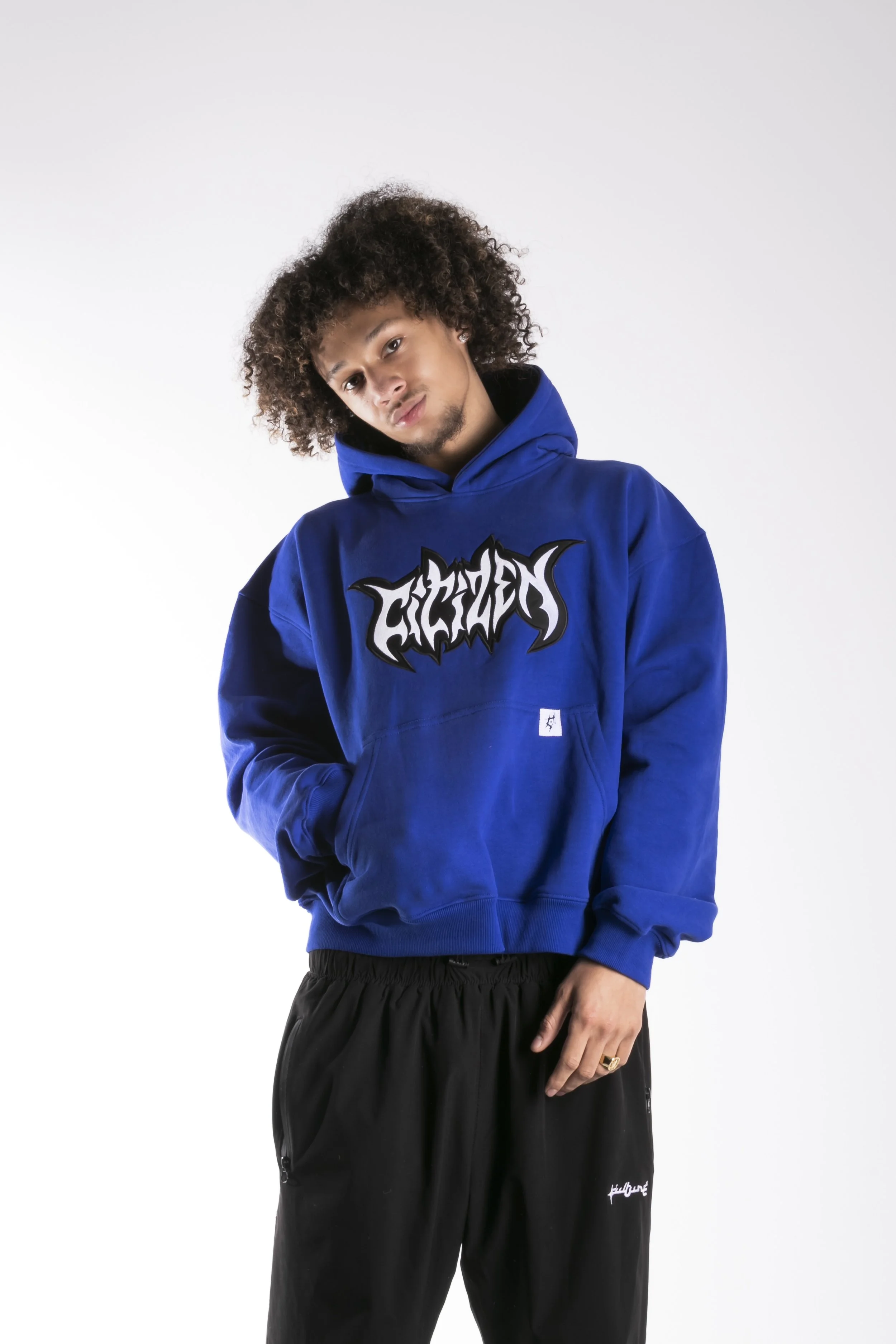 Jeune homme portant un sweat à capuche bleu avec un logo noir et blanc, et un pantalon noir, posant contre un fond blanc.