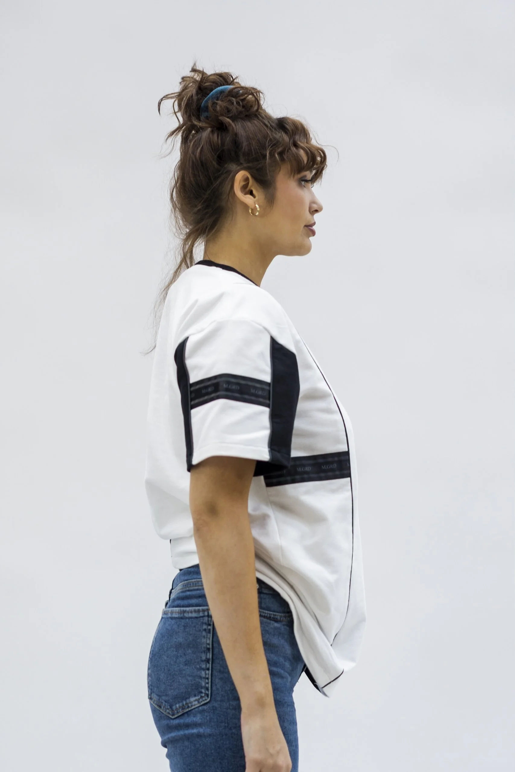 Femme en jean et chemise blanche vue de profil contre un mur blanc.