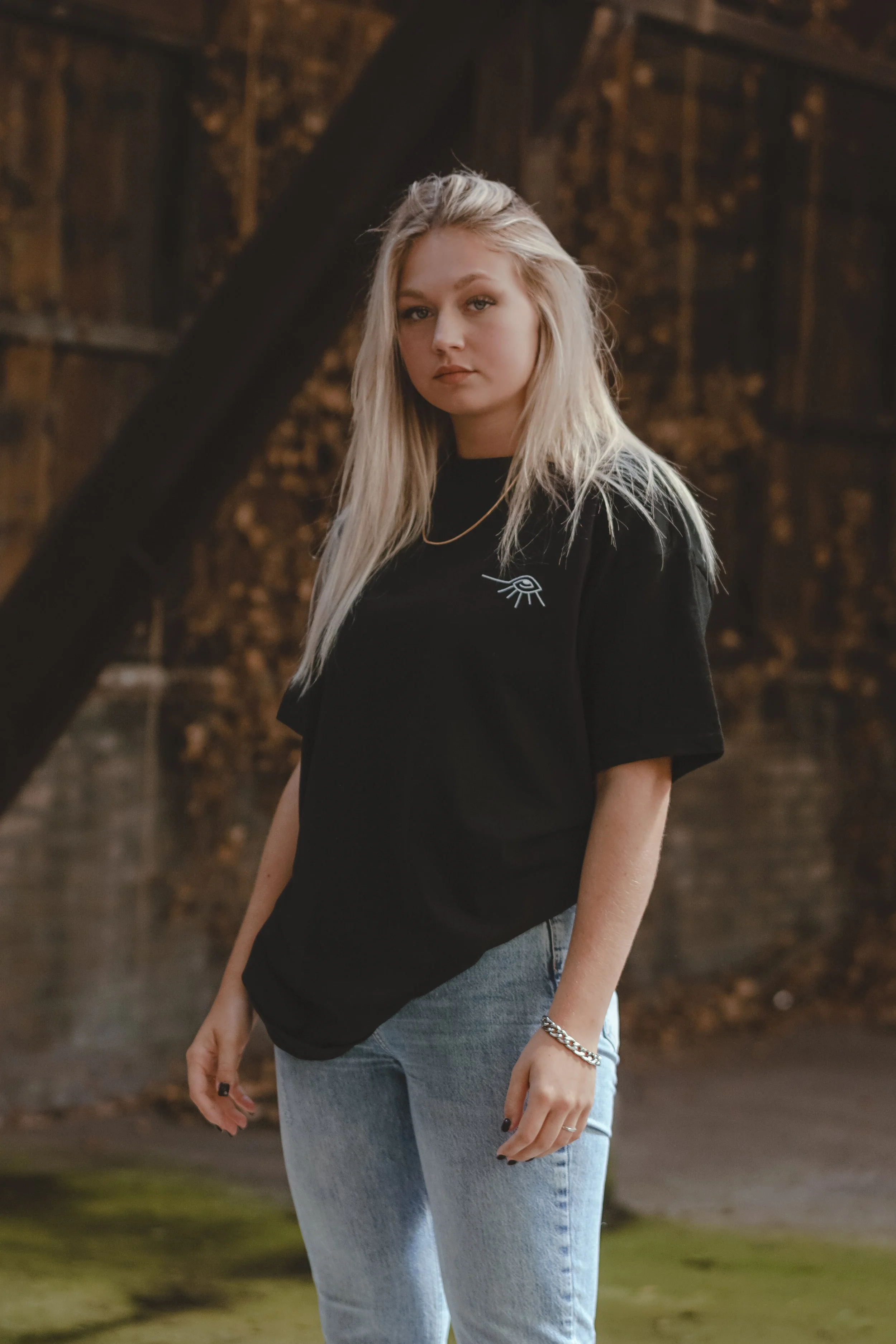 Une jeune femme aux cheveux blonds, portant un t-shirt noir avec un dessin d'œil, et un collier doré, dans un environnement intérieur en bois.