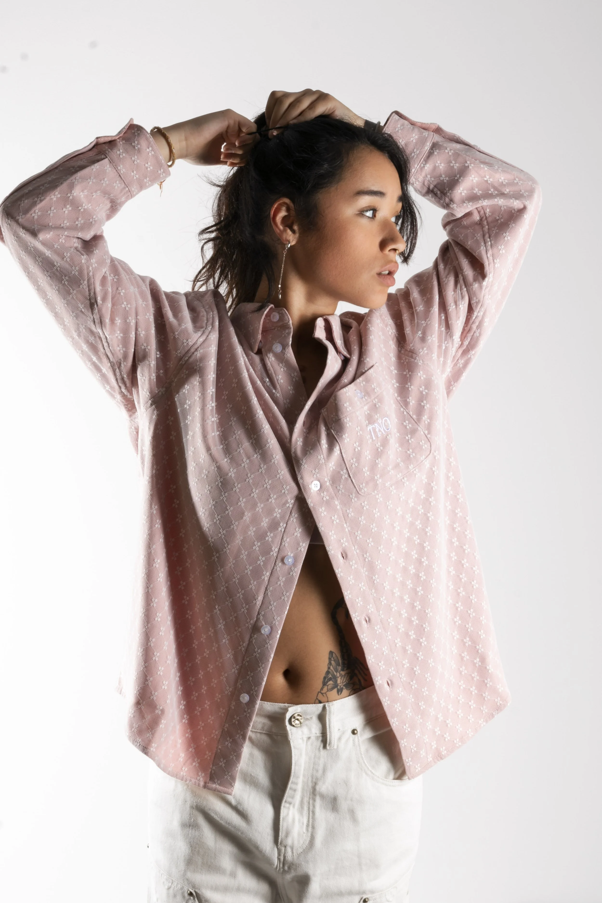 Jeune femme avec une chemise rose clair et un tatouage visible à l'abdomen, levant ses bras pour attacher ses cheveux, en fond blanc.