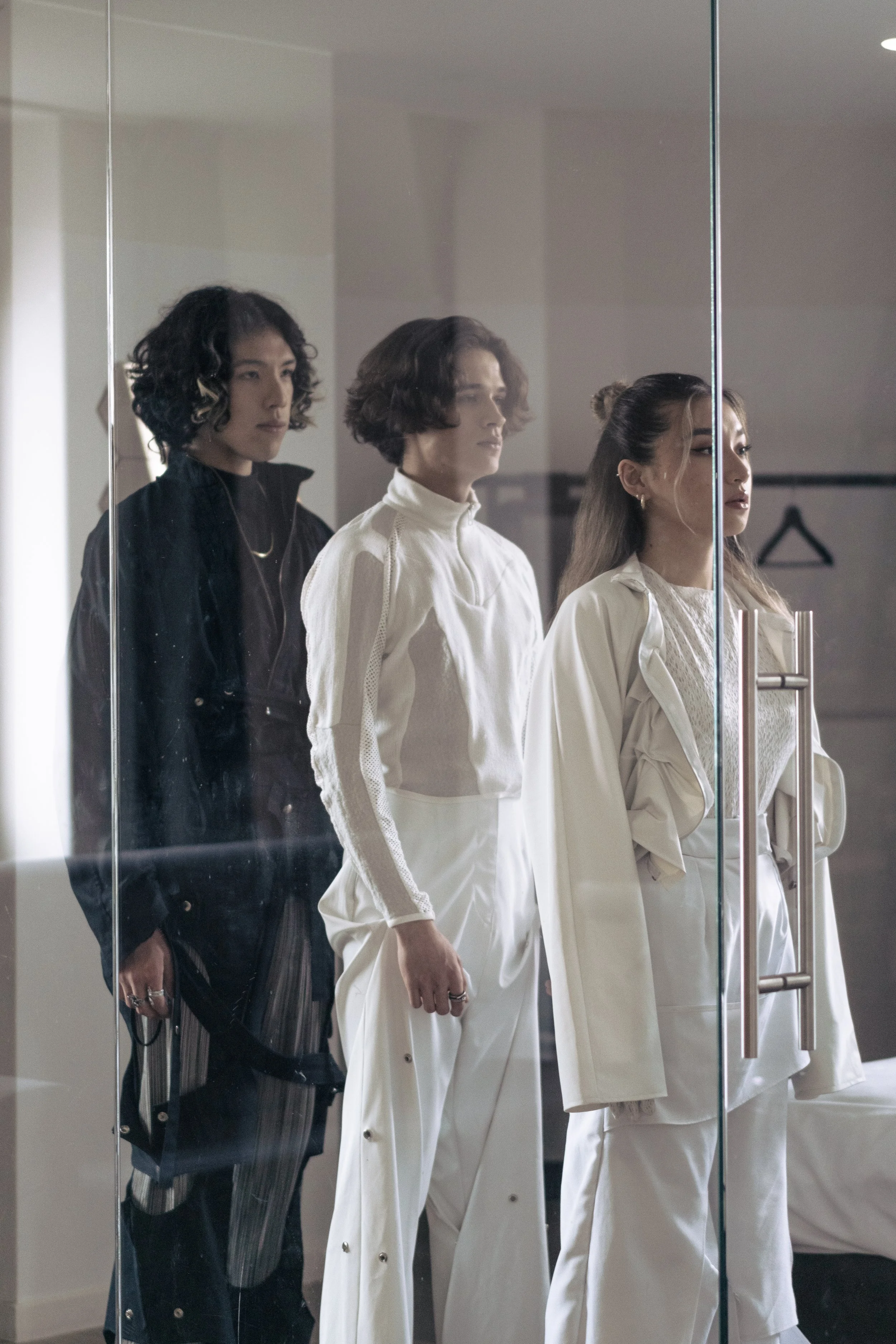 Trois femmes regardant dans un miroir en verre dans un intérieur moderne. La femme à gauche porte une veste noire avec rayures verticales. La femme au centre porte un vêtement blanc avec des détails en maille, et la femme à droite porte une tenue bla