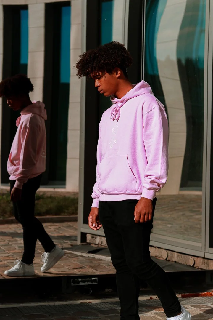 Jeune homme avec cheveux frisés portant un sweat à capuche rose clair, debout devant une surface miroitante, avec une réflexion partielle de lui-même visible.