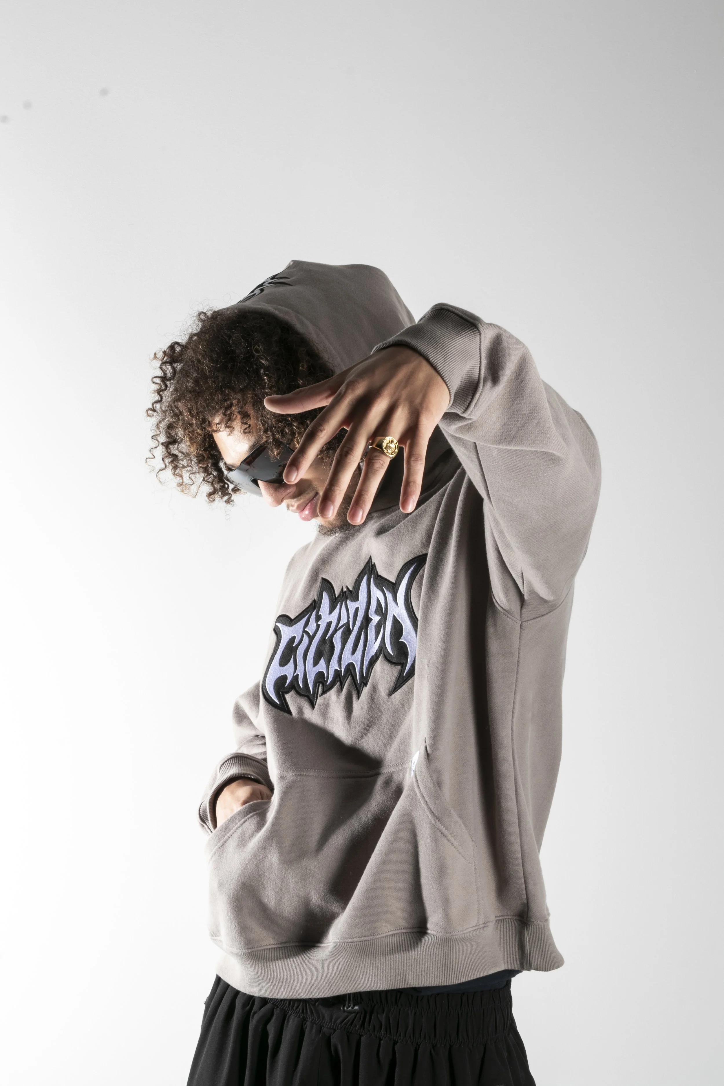 Jeune homme portant un hoodie gris avec un logo stylisé, portant des lunettes de soleil, avec une main proche du visage portant une bague en or, en position décontractée sur un fond blanc.