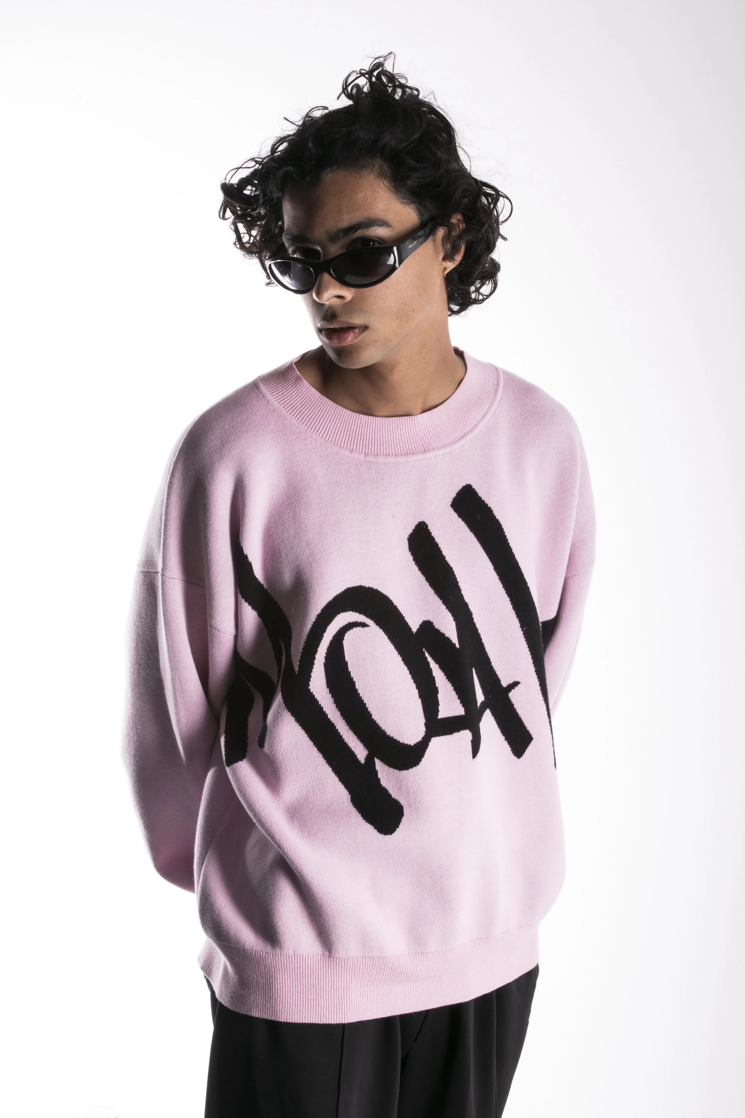 Jeune homme portant un sweat-shirt rose avec un design graphique noir, des lunettes de soleil noires, cheveux bouclés noirs, posé contre un fond blanc.