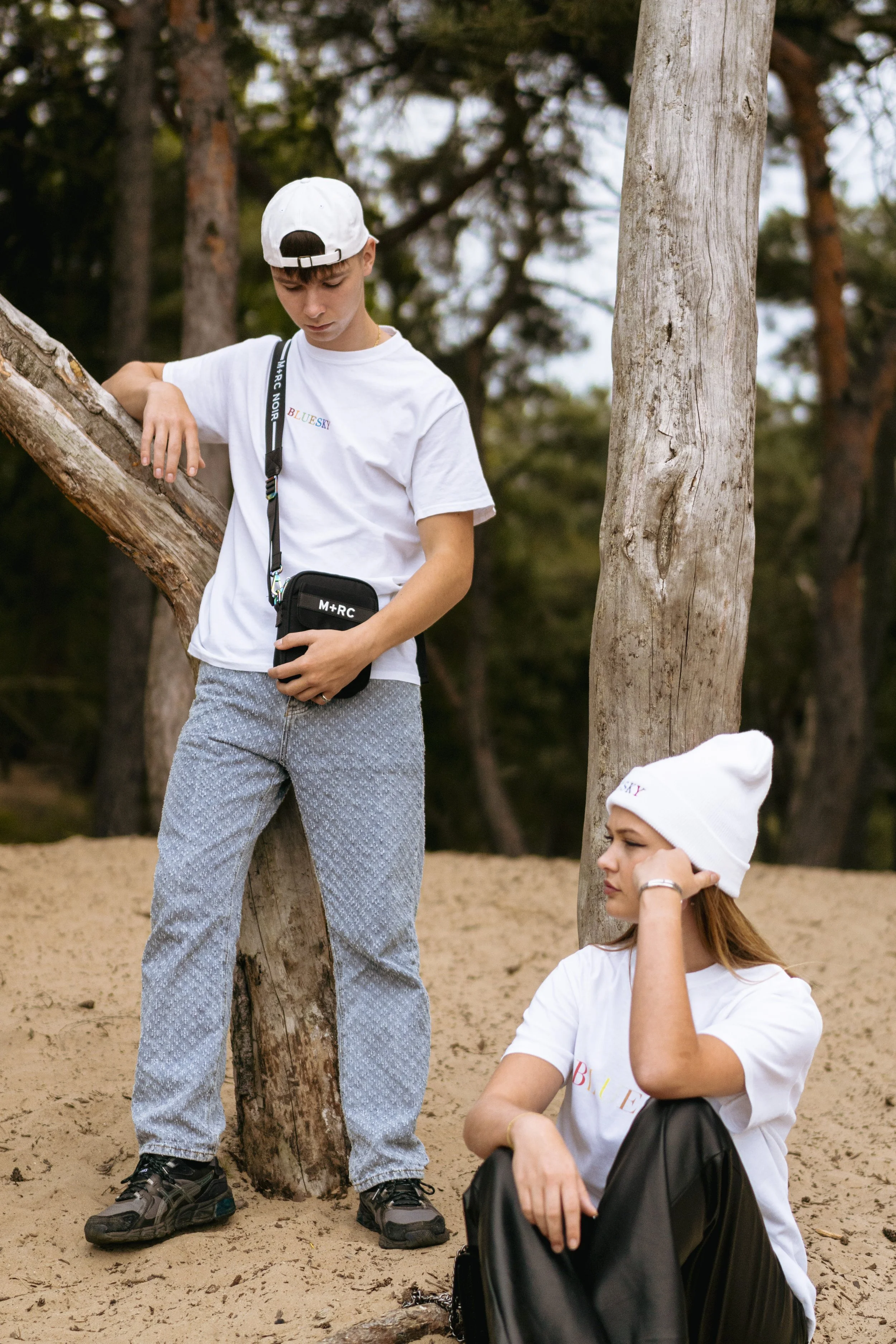Un jeune homme et une jeune femme dans une forêt, l'homme debout en regardant vers le bas et la femme assise sur le sable, portant un bonnet blanc, t-shirts blancs, et le jeune homme porte un sac à bandoulière.