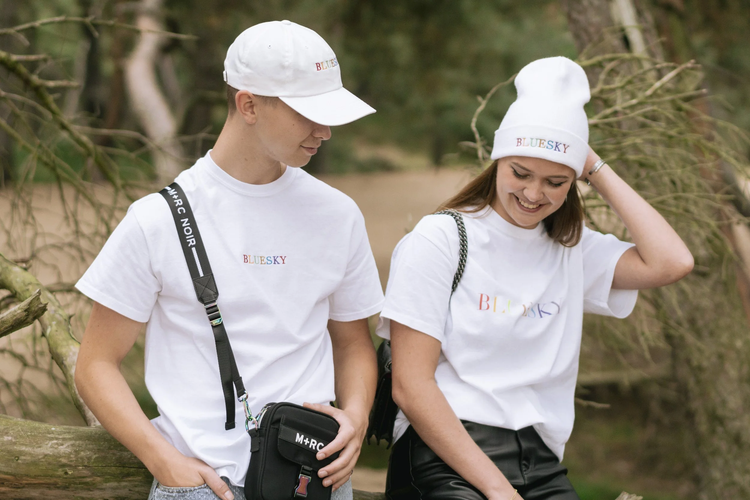 Un jeune homme et une jeune femme souriants, portant des vêtements blancs avec le logo "BLUESKY", marchent dans une forêt de conifères, vêtus de t-shirts et de chapeaux blancs avec le logo en couleur.