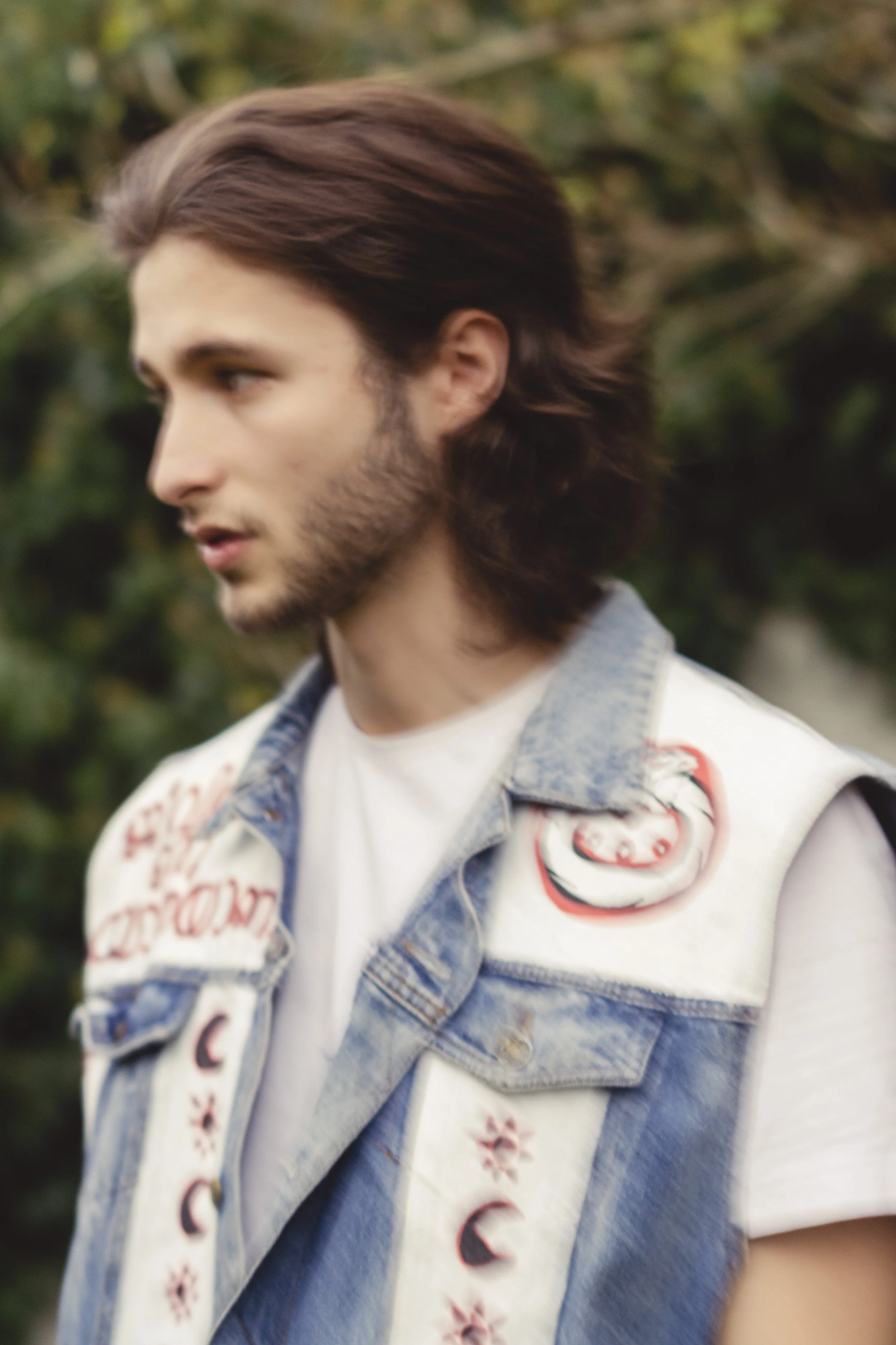 Un jeune homme avec des cheveux longs et une barbe, portant une veste en jean avec des patches, se tient à l'extérieur avec un fond de verdure.