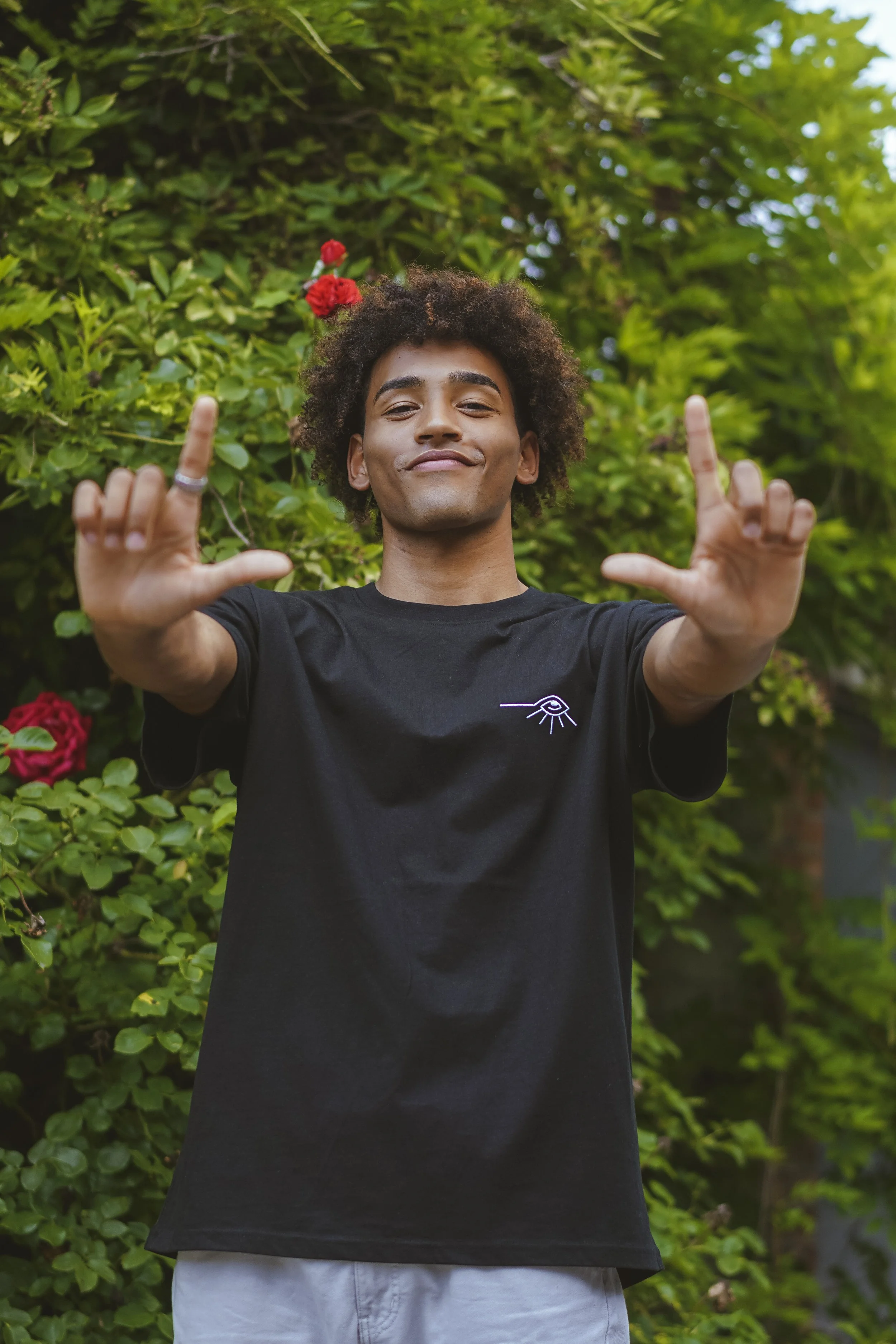 Un jeune homme souriant dans un jardin, portant un t-shirt noir avec un dessin d'œil simple, faisant un geste de la main avec les doigts en L.