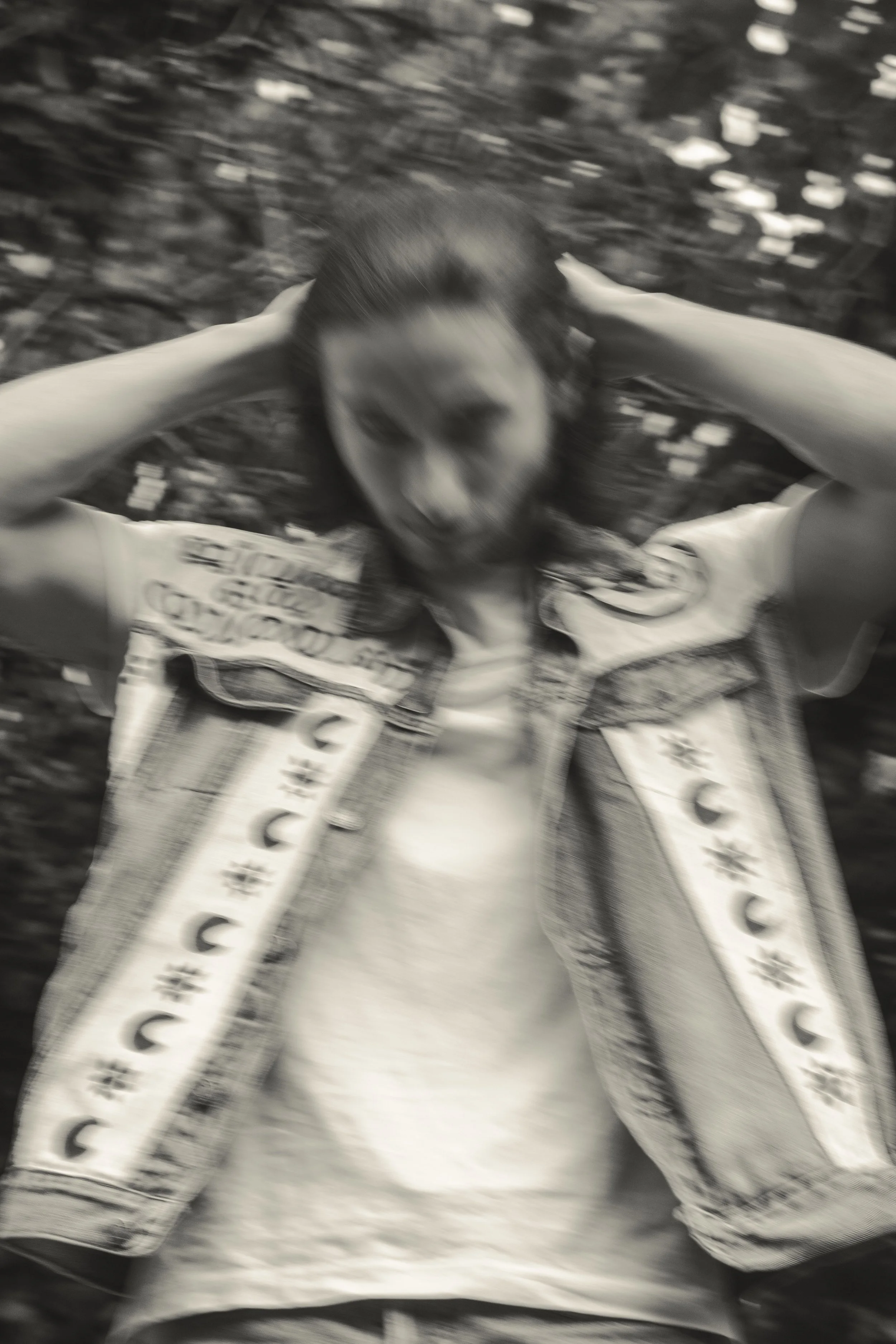 Femme souriante avec les mains derrière la tête, vêtue d'une veste en jean, dans un environnement naturel, photographiée en noir et blanc avec un effet de flou.