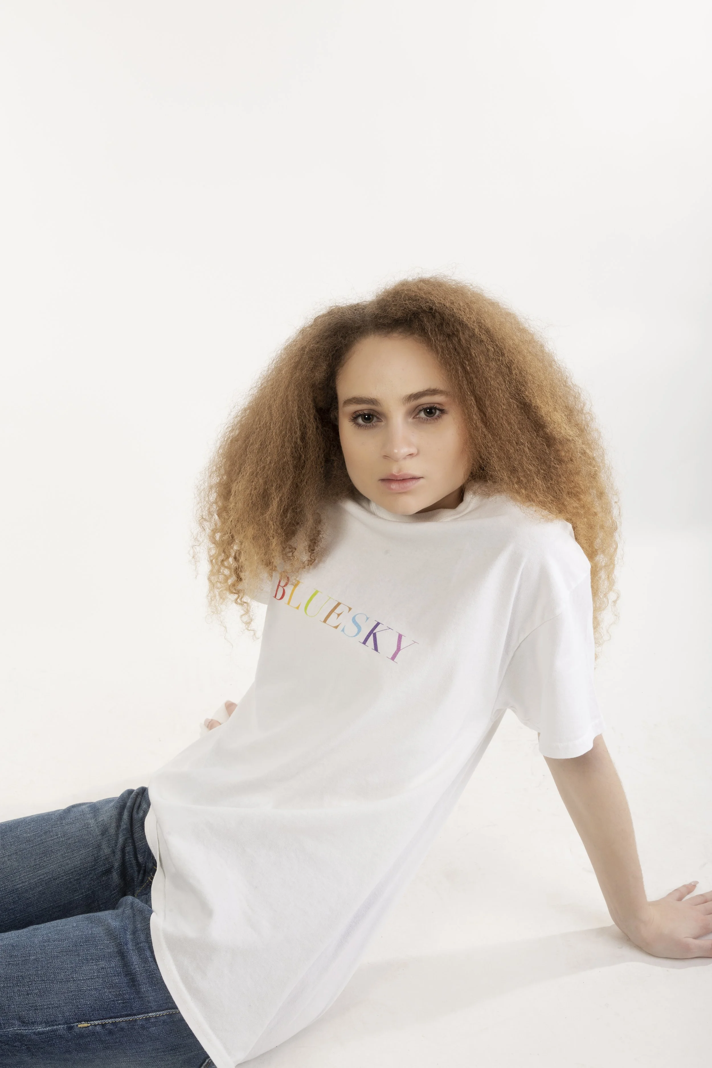 Jeune femme aux cheveux bouclés, portant un t-shirt blanc avec le mot 'BLUESKY' écrit en couleurs arc-en-ciel, assise sur un fond blanc.