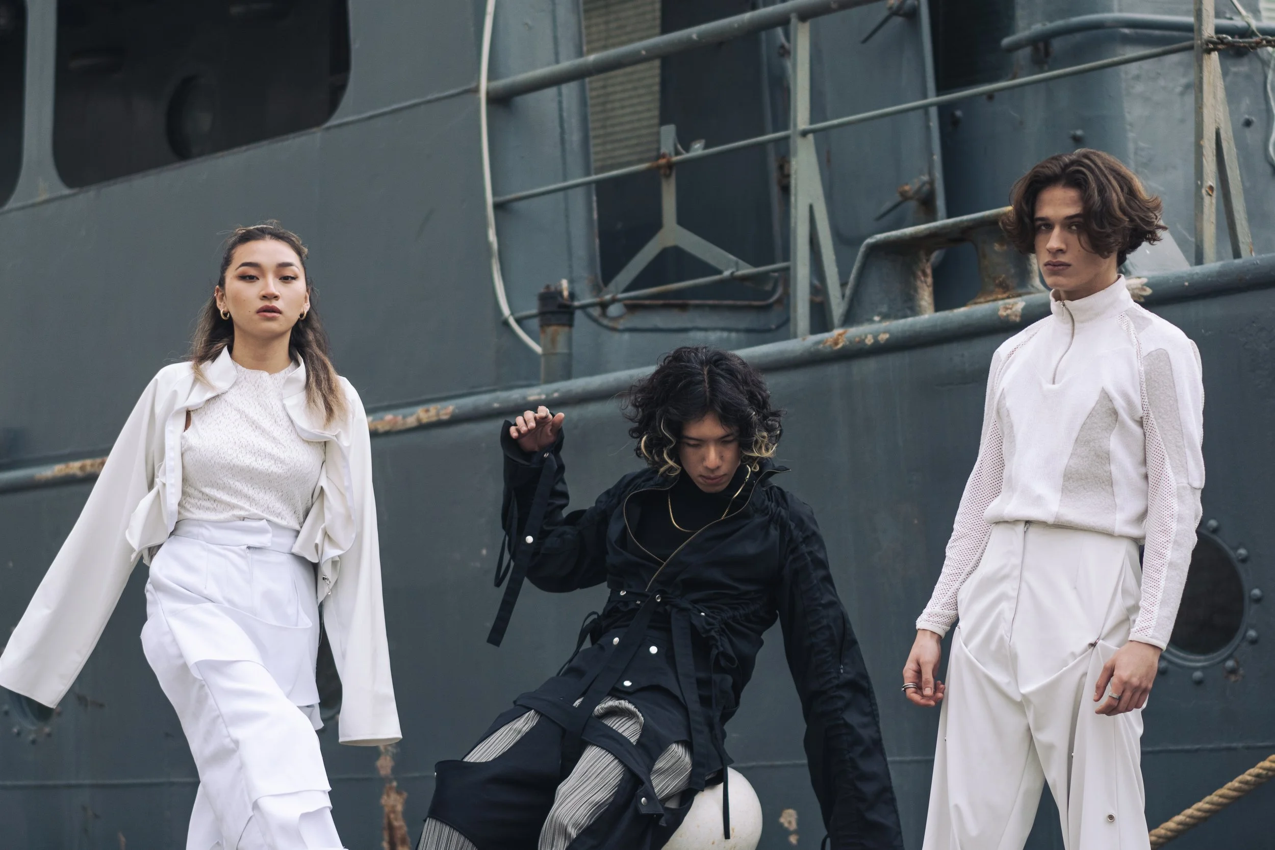 Trois jeunes personnes posent devant un navire, portant des vêtements modernes et stylés.