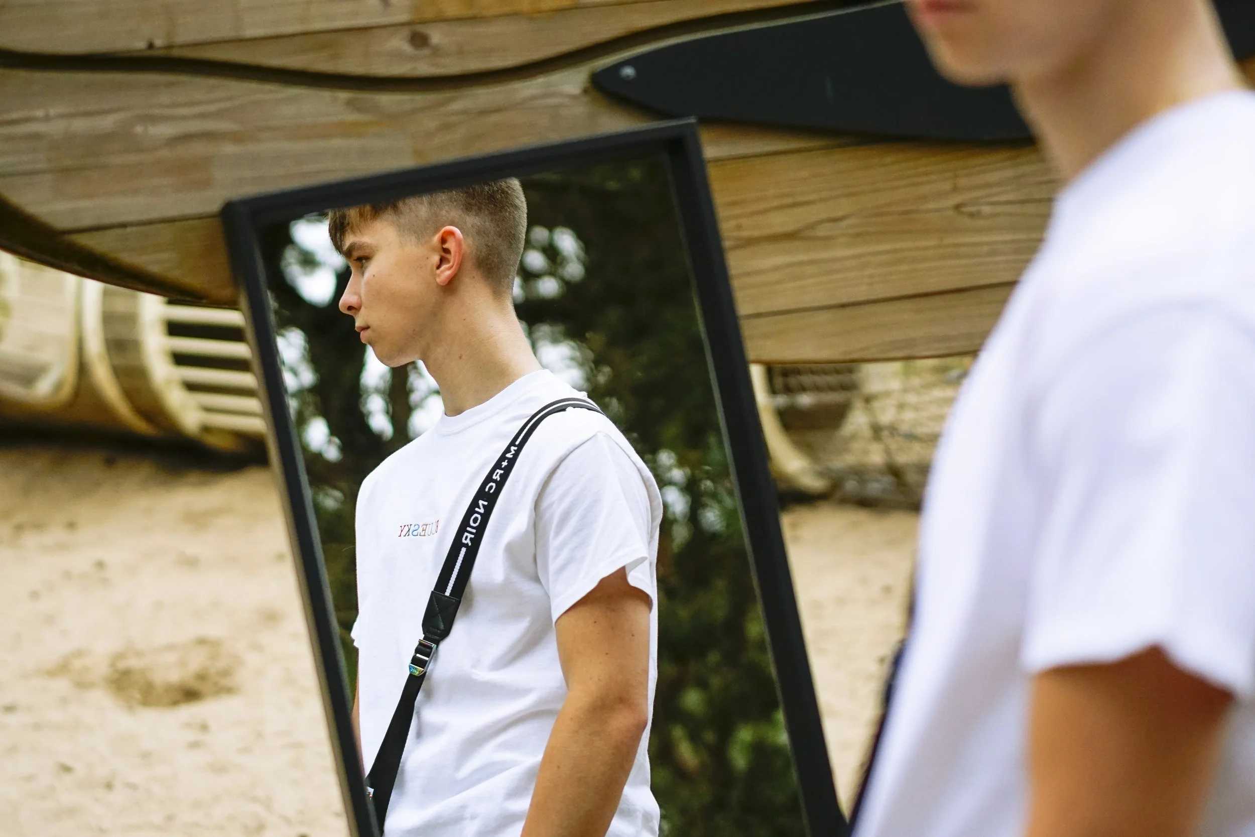 Jeune homme observant son reflet dans un miroir en plein air, portant un t-shirt blanc et un sac à bandoulière, avec un fond naturel.