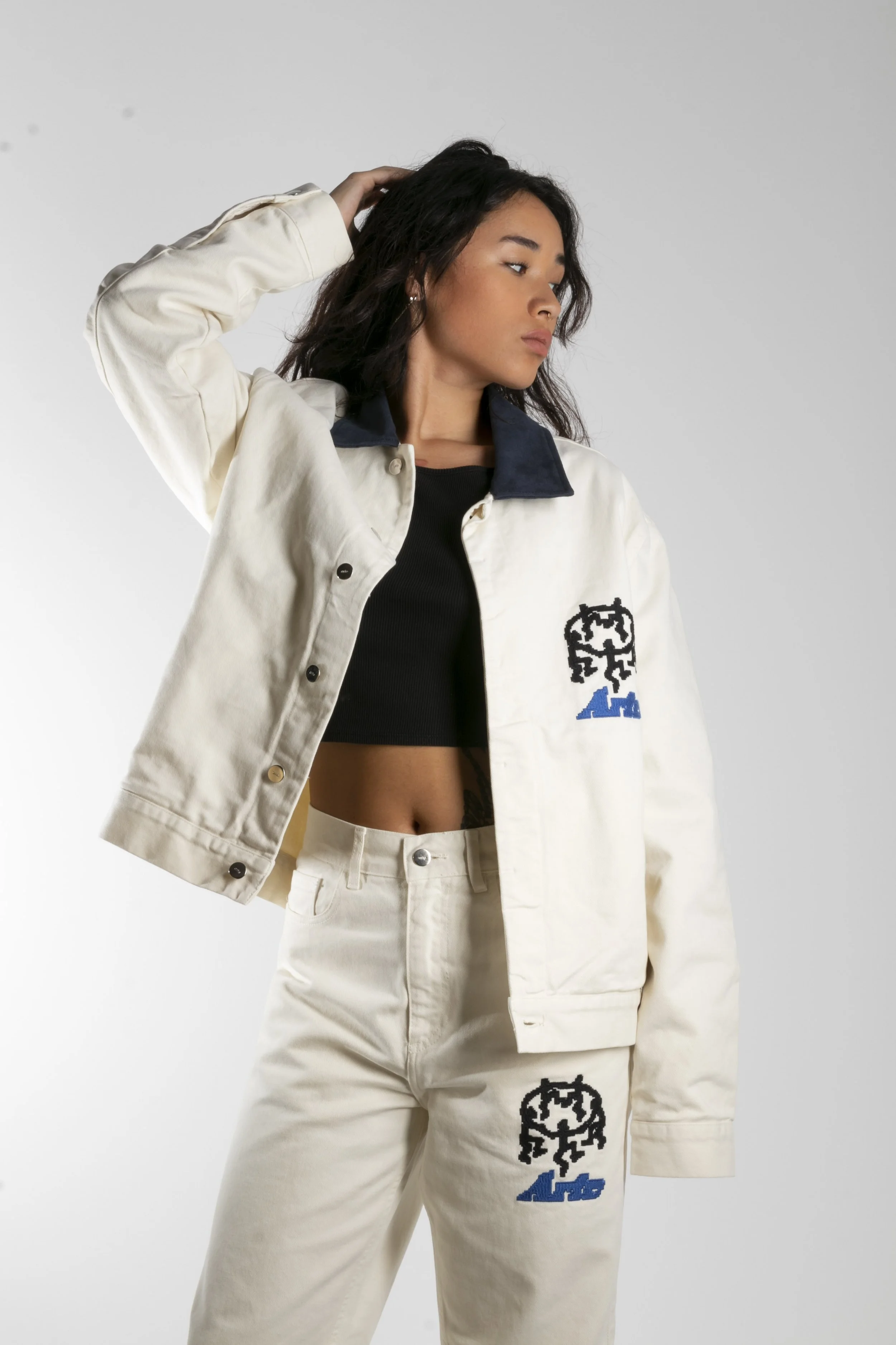 Jeune femme portant une veste blanche, un crop top noir et un pantalon blanc avec des logos noirs et bleus.