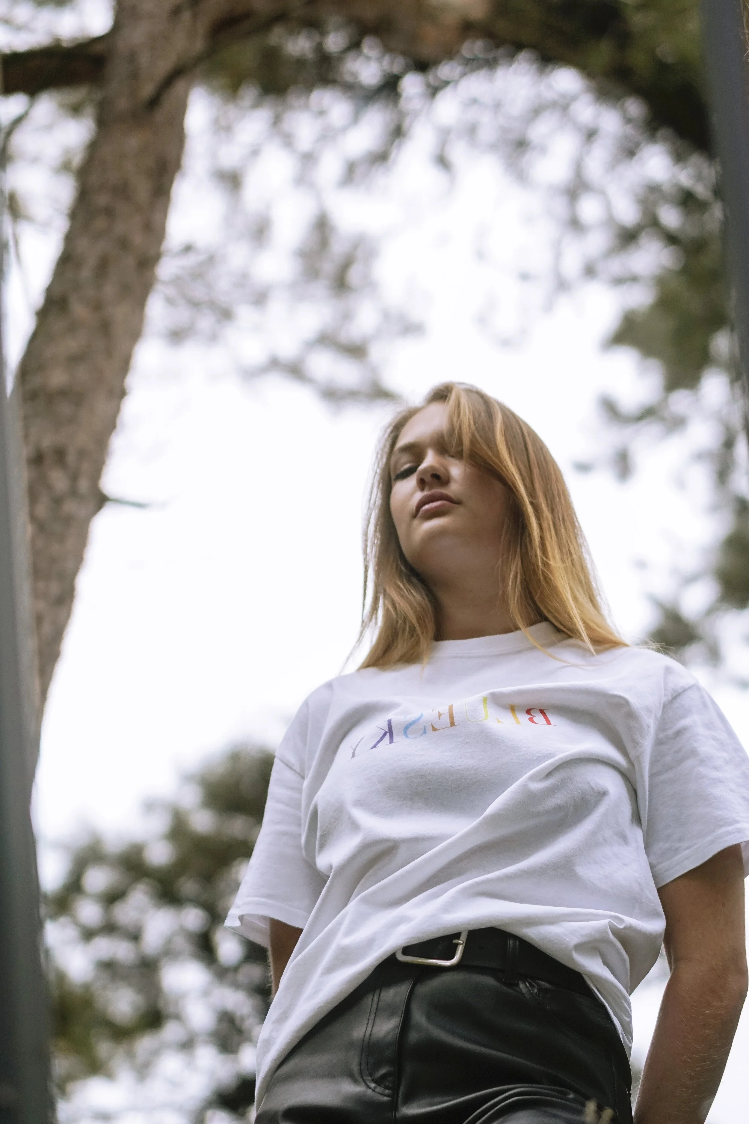 Jeune femme aux cheveux roux portant un t-shirt blanc avec des lettres colorées, vue de bas vers haut, arbres en arrière-plan.