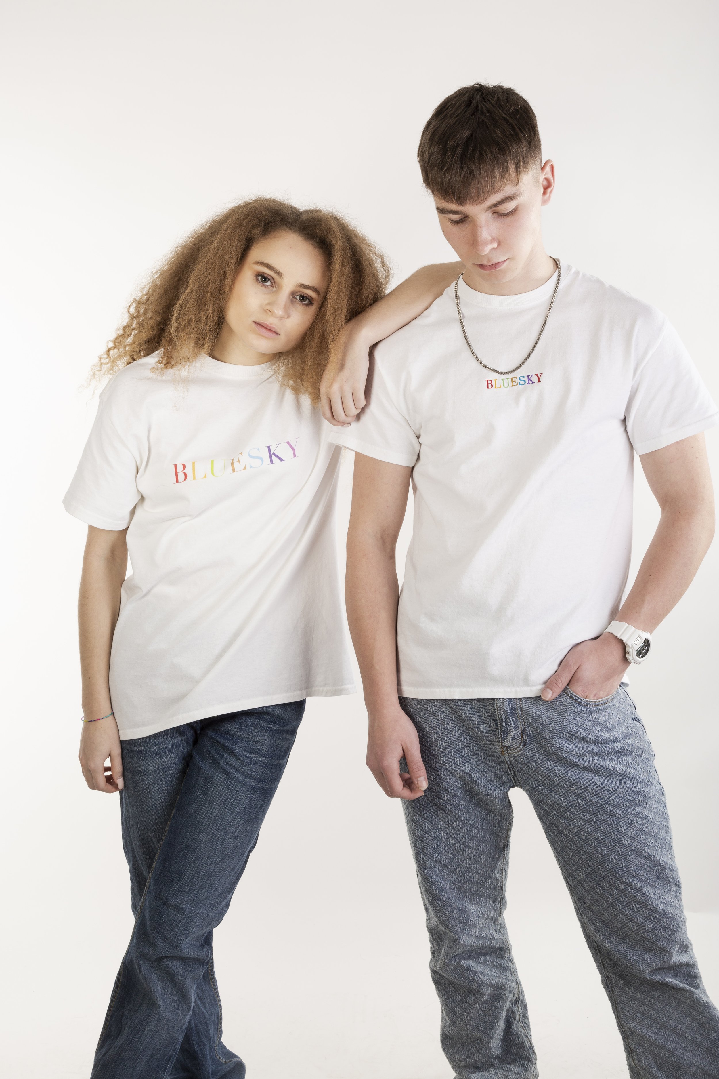 Deux jeunes portant des t-shirts blanc avec le mot 'BLUESKY' écrit en lettres colorées, pose décontractée contre un fond blanc.