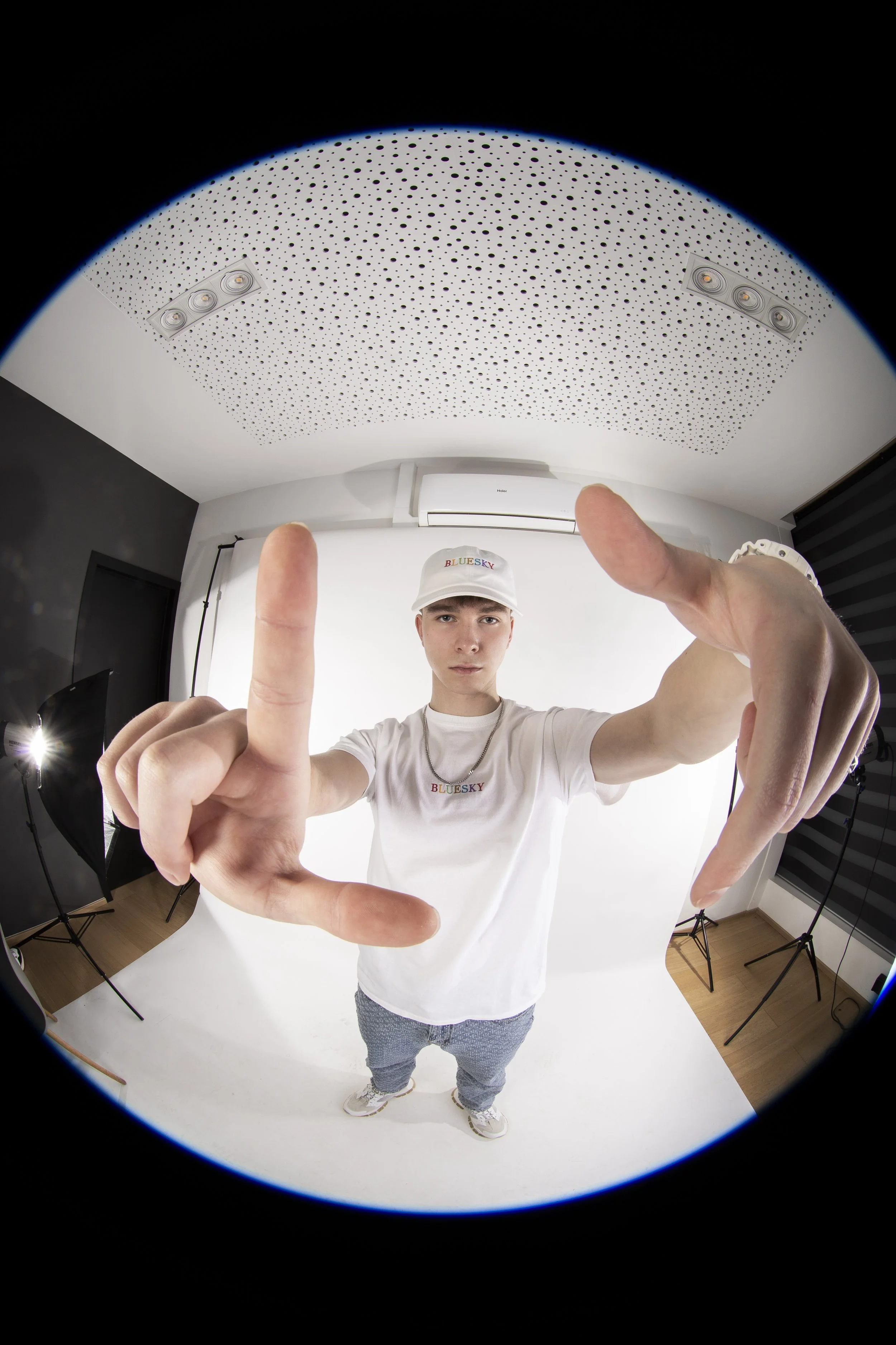 Jeune homme en studio portant un t-shirt et une casquette blancs, faisant un geste de la main vers l'appareil photo avec un fond blanc. Studio équipé de lumières, un fond noir et un plafond acoustique perforé.