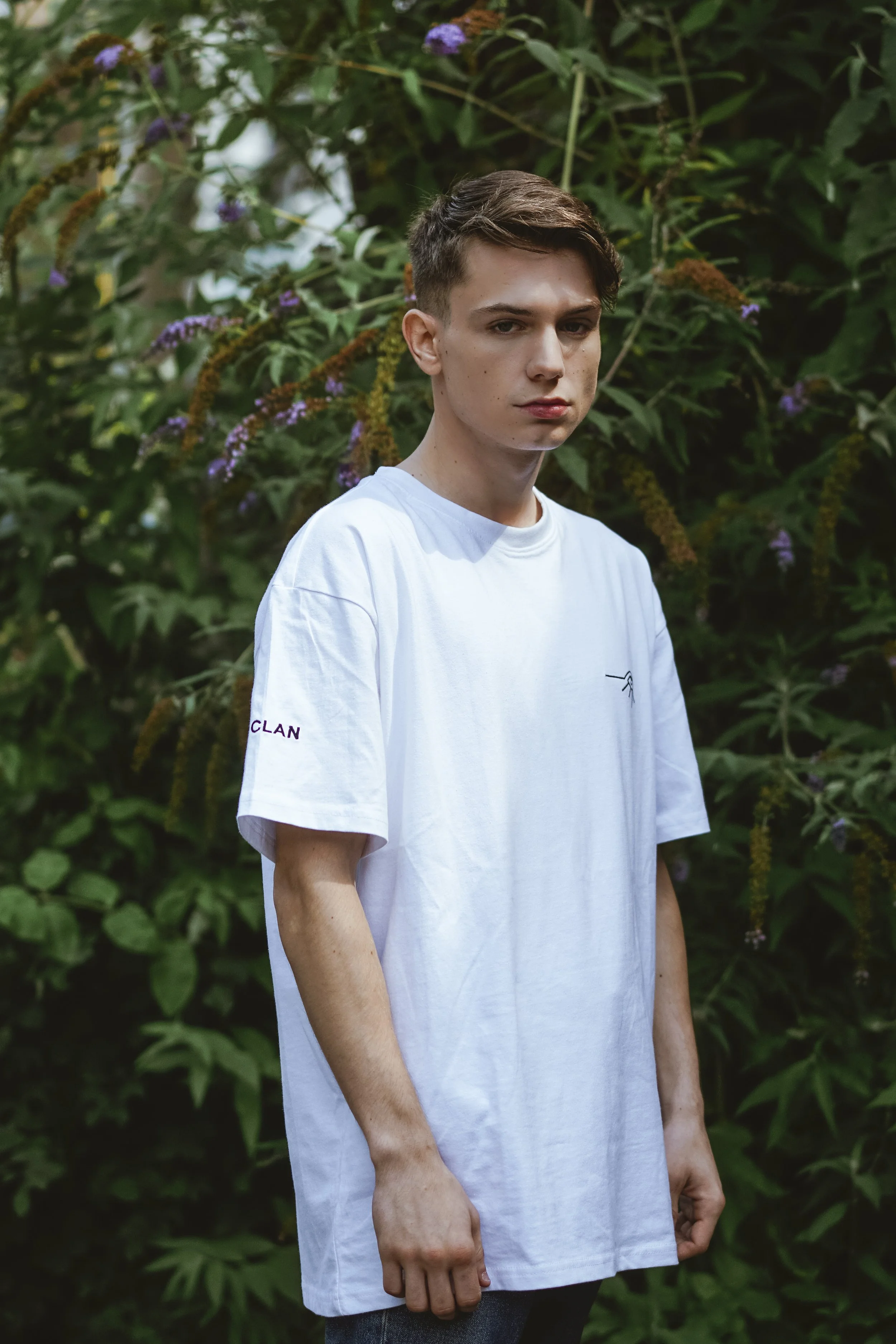 Jeune homme portant un t-shirt blanc, debout devant une végétation luxuriante avec des fleurs violettes et vertes.