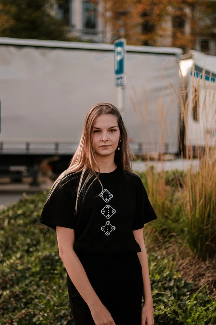 Jeune femme avec des cheveux longs et un t-shirt noir, debout devant un fond de végétation et de camions, dans un environnement urbain