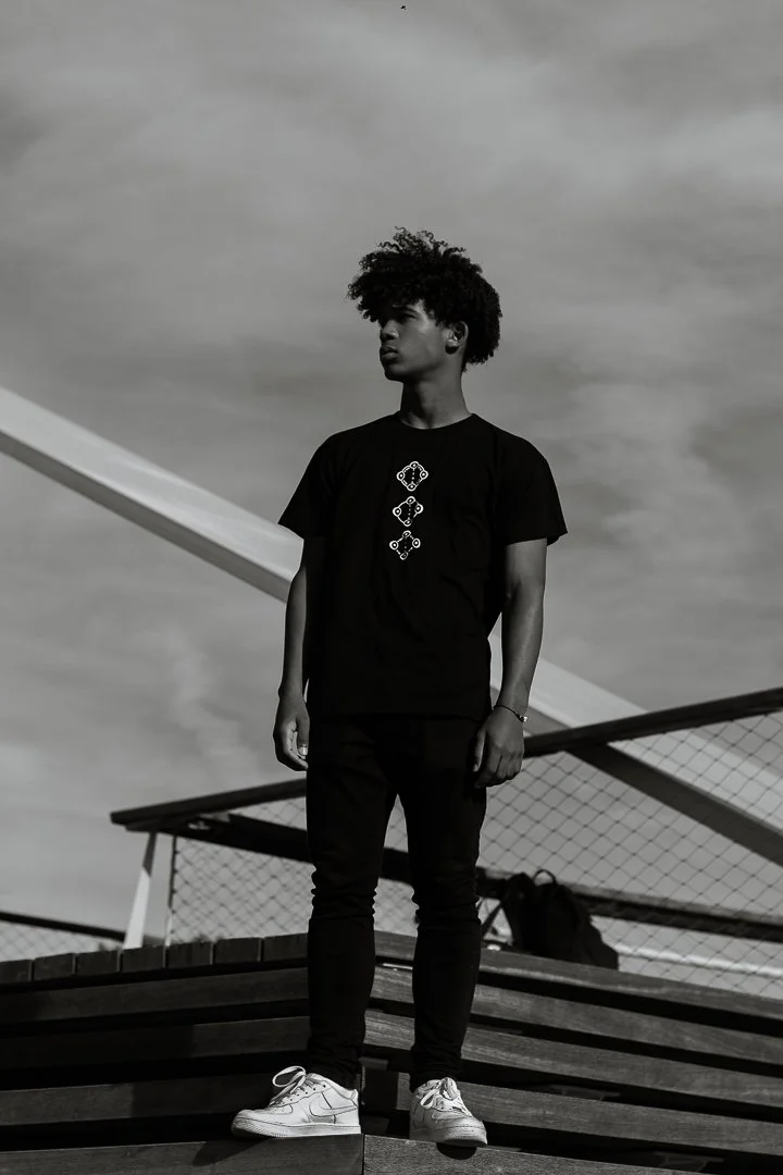 Jeune homme debout sur une plateforme en bois, portant un t-shirt noir avec un motif géométrique, pantalons noirs et des chaussures blanches, dans un environnement extérieur avec un ciel nuageux.