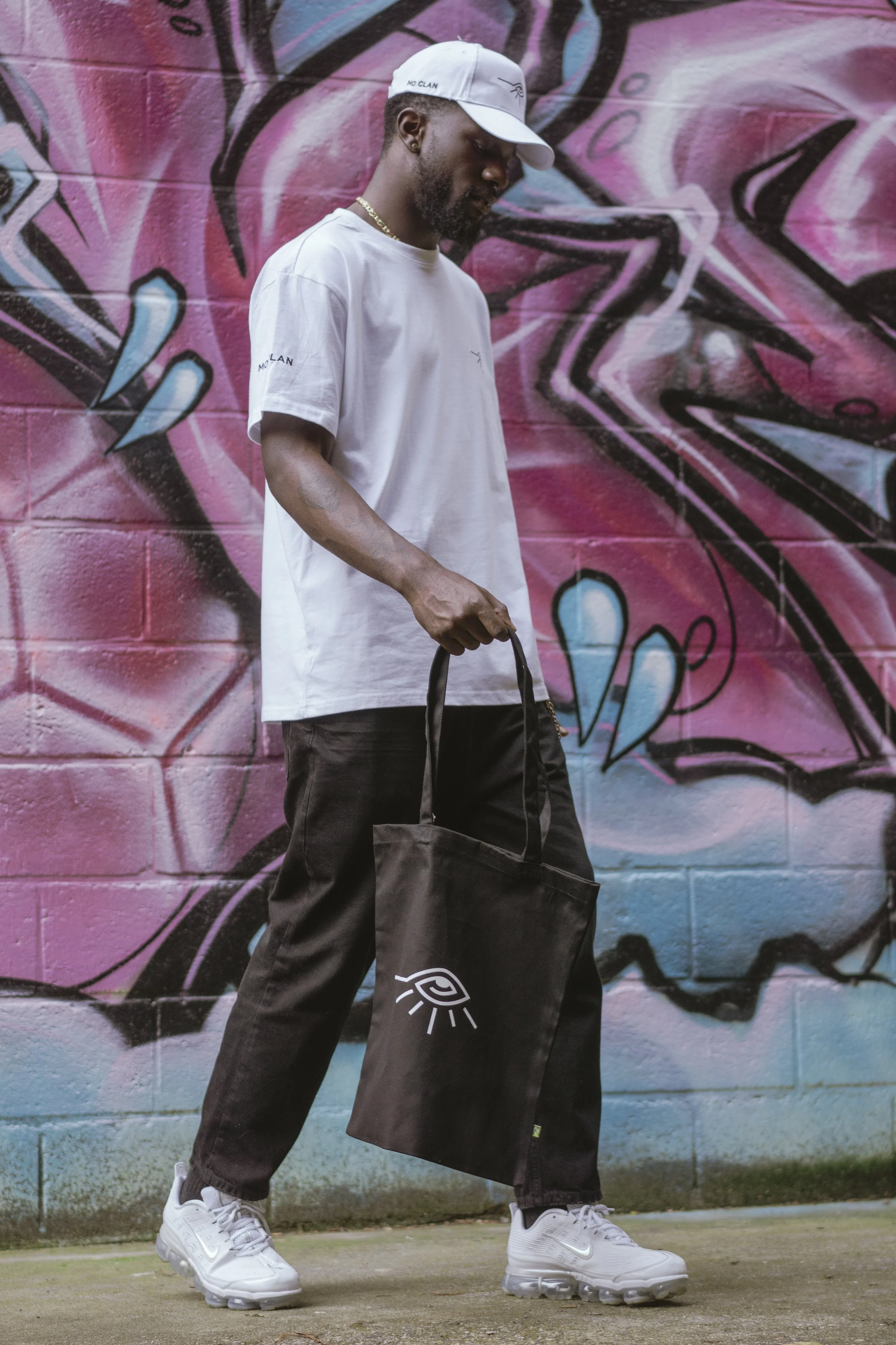 Un homme portant un chapeau blanc, un t-shirt blanc et un pantalon sombre marche devant un mural graffité rose, bleu et noir, tout en tenant un sac noir avec un symbole graphique blanc.