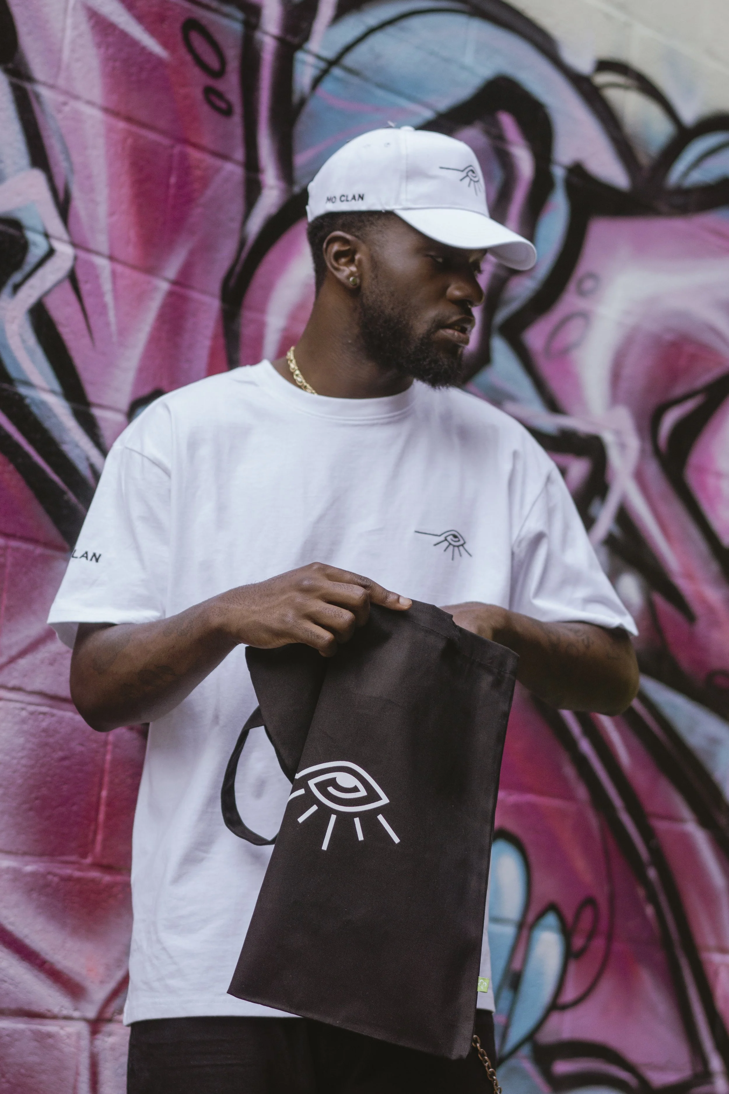 Un homme portant une casquette blanche, un t-shirt blanc avec un logo d'œil stylisé, tient un sac noir avec un logo d'œil stylisé, se tenant devant un mur de graffitis colorés.