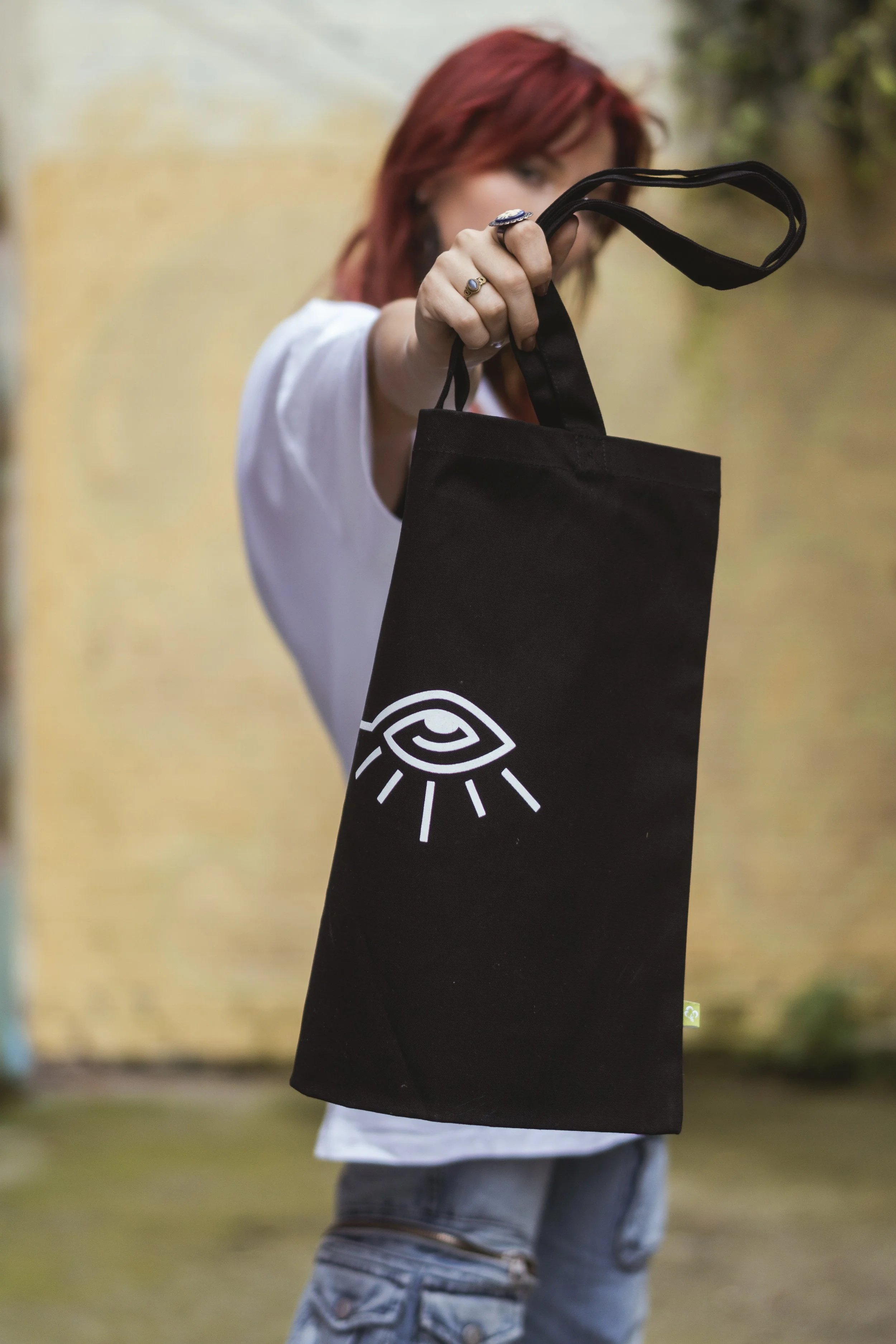 Femme aux cheveux roux tenant un sac en tissu noir avec un logo blanc, portant une chemise blanche et un jean, dans un environnement extérieur.