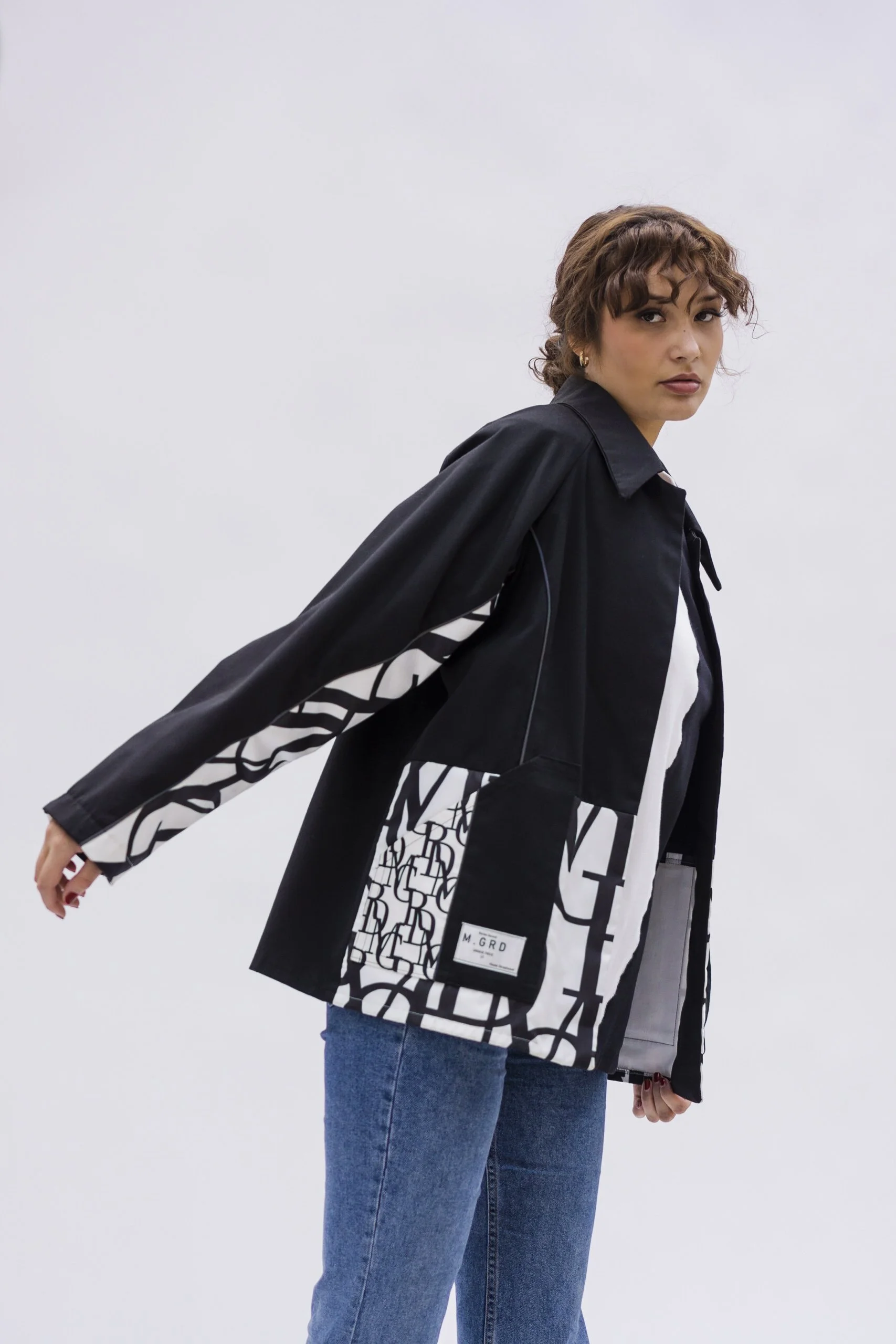 Femme portant une veste noire et un jean bleu clair, debout contre un fond blanc.