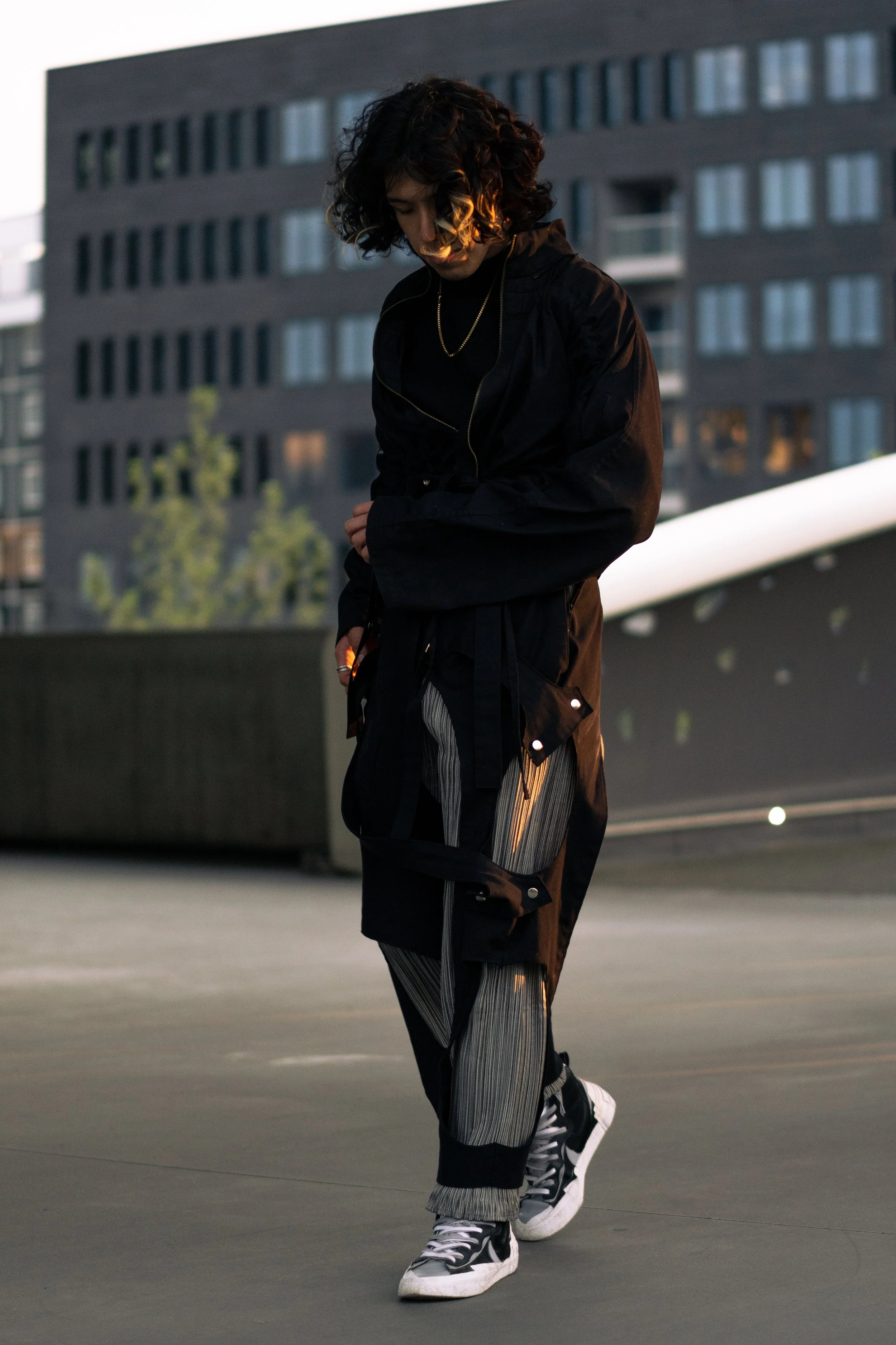 Une jeune femme en vêtements décontractés, avec un sweat noir, un pantalon à rayures gris et noires, et des baskets, marche dans un environnement urbain lors du coucher du soleil.