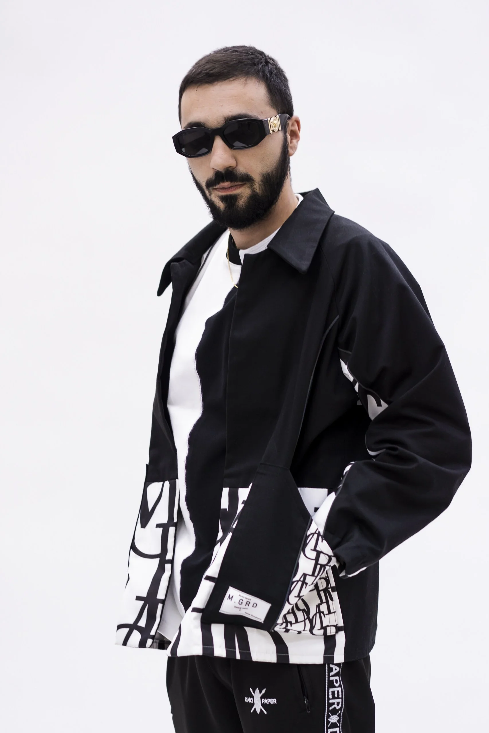 Homme portant des lunettes de soleil noires, une veste noire avec des motifs noirs et blancs, et un pantalon noir dans un fond blanc.