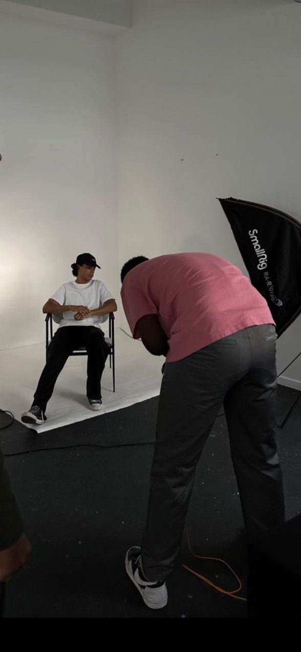 Une séance photo en studio avec une personne assise sur une chaise, et un photographe prenant la photo. La scène est simple et le fond est blanc.