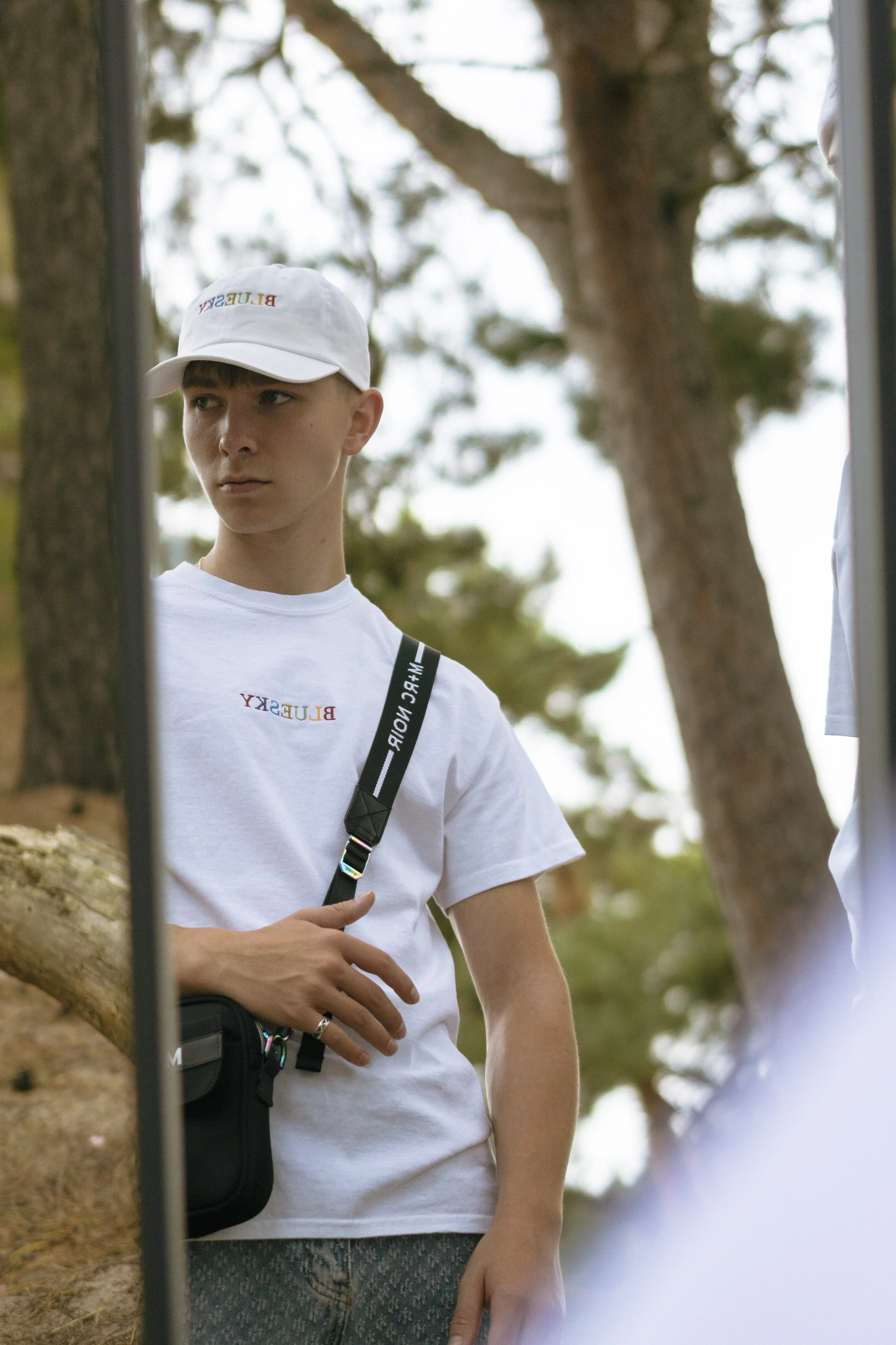 Un jeune homme portant un t-shirt blanc avec le logo BLUESKY, un sac à bandoulière, une casquette blanche avec le même logo, regardant pensivement à travers un miroir en plein air, avec des arbres en arrière-plan.
