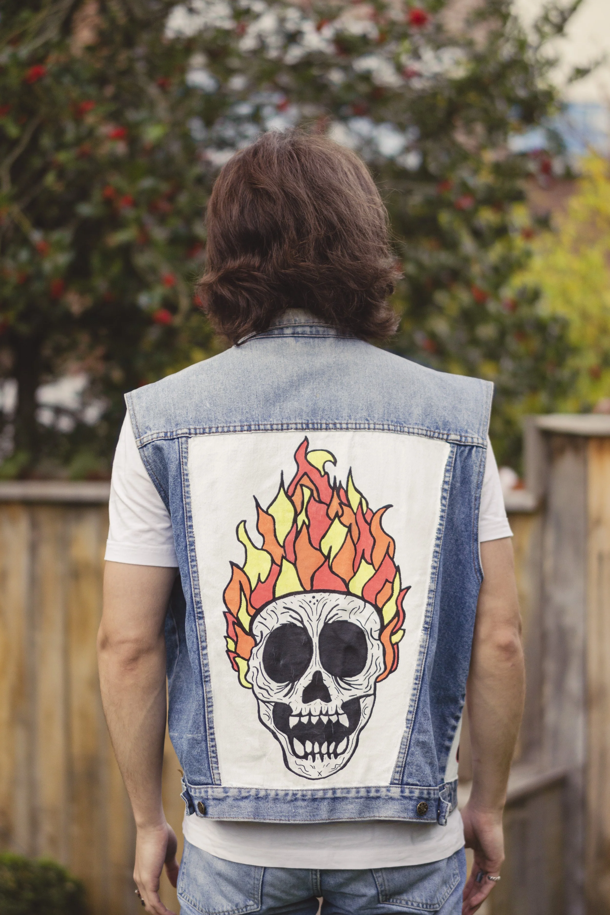 Person vue de dos portant un gilet en jean avec un dessin de crâne en feu sur le dos, dans un jardin avec un arbre à fleurs en arrière-plan.
