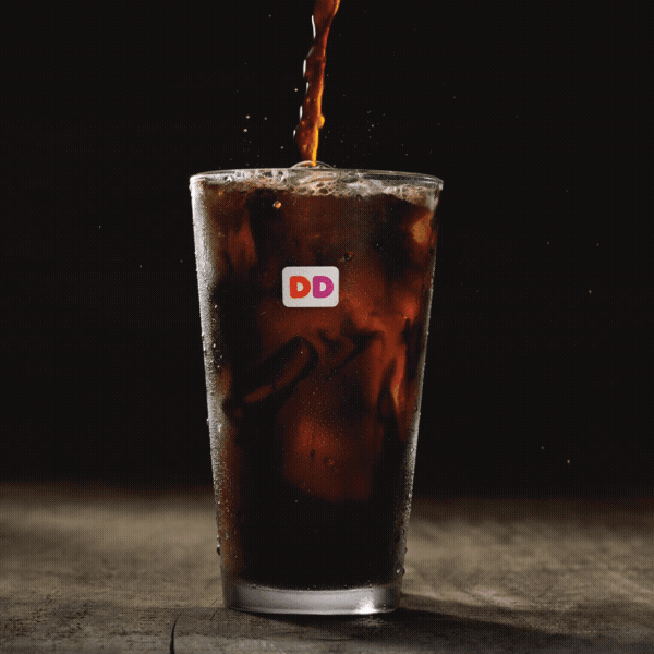 A glass of iced coffee with ice being filled from above, featuring the Dunkin Donuts logo on the glass.