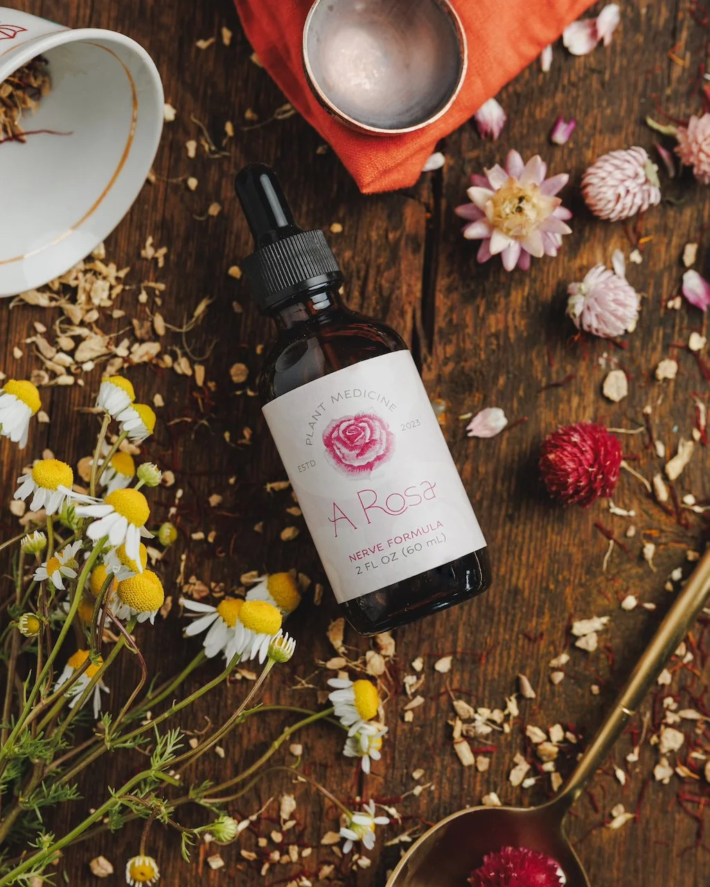 A dark amber bottle labeled "A Rosa" with a pink rose illustration, laying on a wooden surface surrounded by flowers, petals, a white dish, a metallic cup, a red cloth, and a gold spoon.